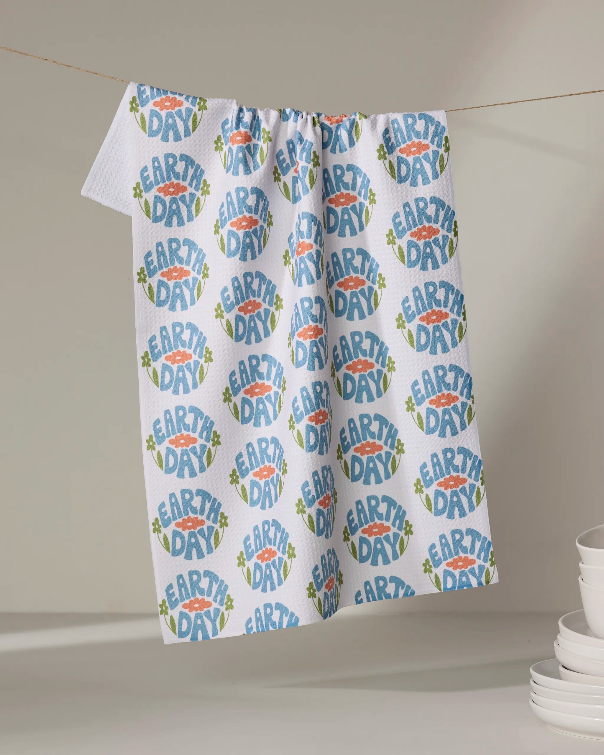 A white, ultra-absorbent Kayla Ann Earth Day Stamp tea towel hangs on a line, adorned with blue, green, and orange circular patterns. Stacks of white bowls and plates are arranged on the right side of the table below.