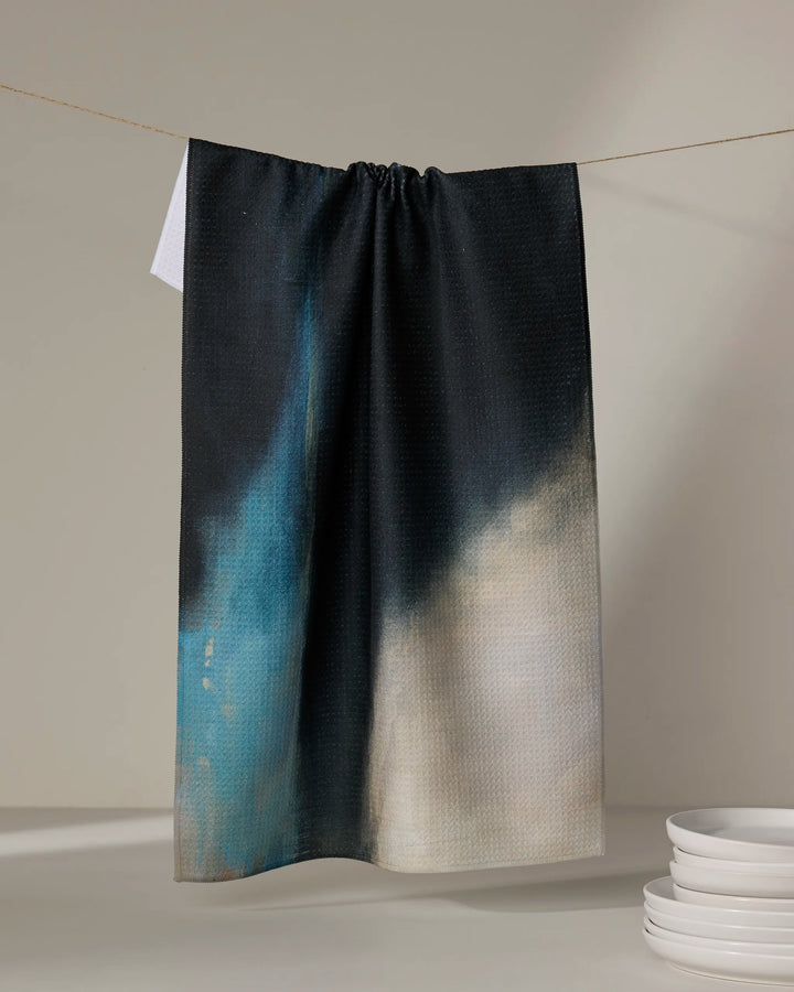 The "Earth Erupted" kitchen towel by Jennifer Janesko, featuring an abstract black, blue, and beige design printed on recycled materials, hangs on a line. White plates are stacked in the lower right corner against a neutral background.