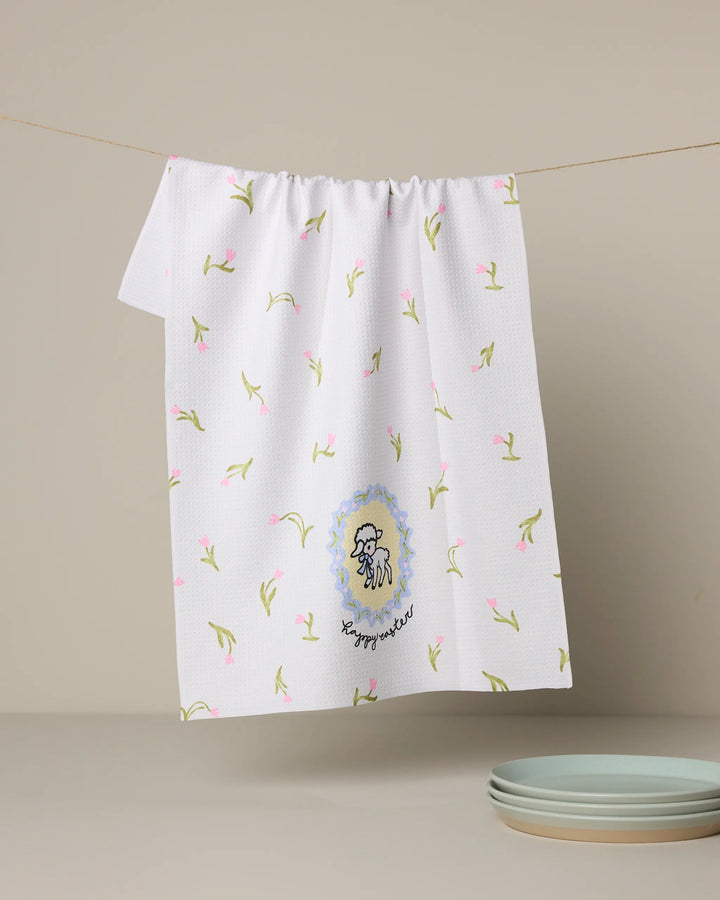 The Easter Little Lamb kitchen towel by Geometry, made from recycled materials, features a cartoon lamb and "happy little" embroidered near the bottom. White with small pink and green flowers, it hangs on a clothesline beside three stacked light blue plates.