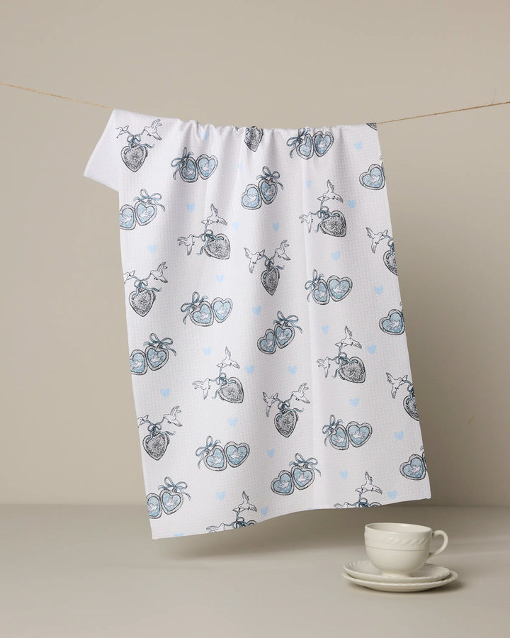 The Geometry “Easter Peace” white Geoweave kitchen tea towel, featuring a pattern of anatomical and blue hearts, hangs on a string above a white teacup and saucer set on a beige surface.