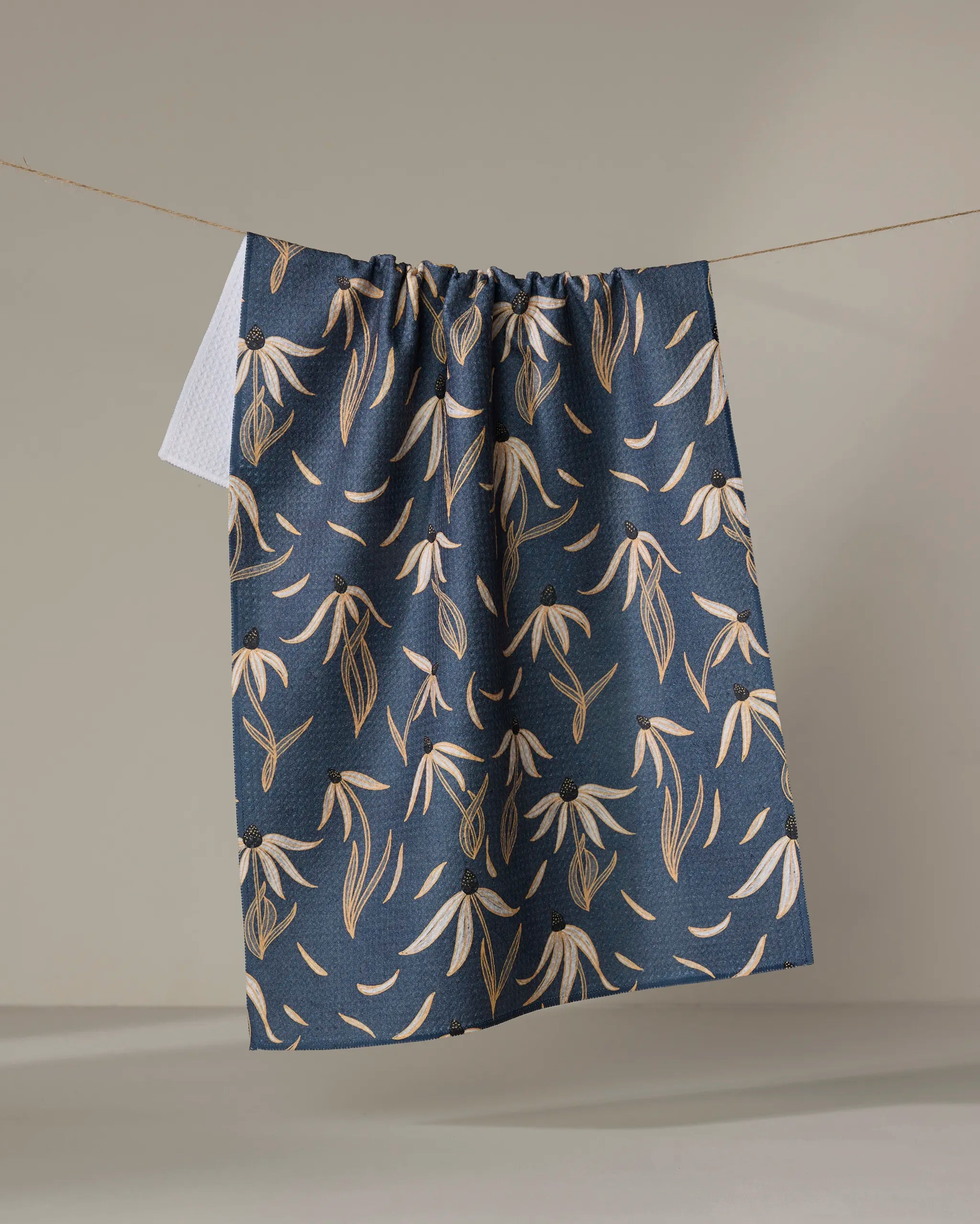 The "Echinacea" kitchen towel by Katie O'Shea, featuring a dark blue background with beige and white floral patterns, hangs on a clothesline with a piece of white textured fabric partially visible behind it.