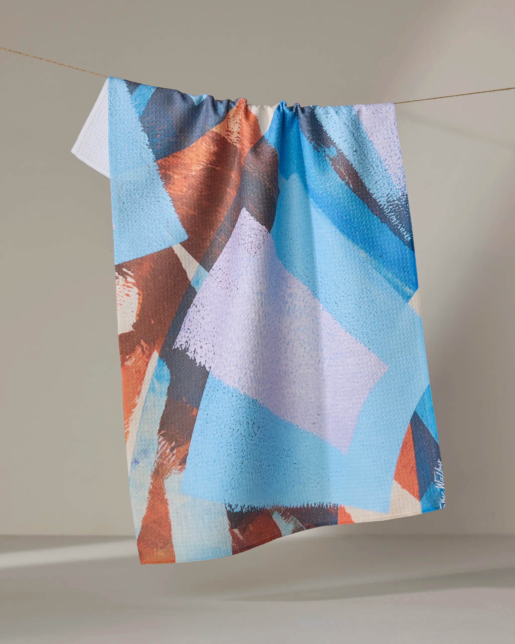 The "Echoes of Color" tea towel by Skye Walker, featuring ultra-absorbent fabric and bold blue, purple, brown, and white geometric shapes, hangs draped over a clothesline against a neutral background.
