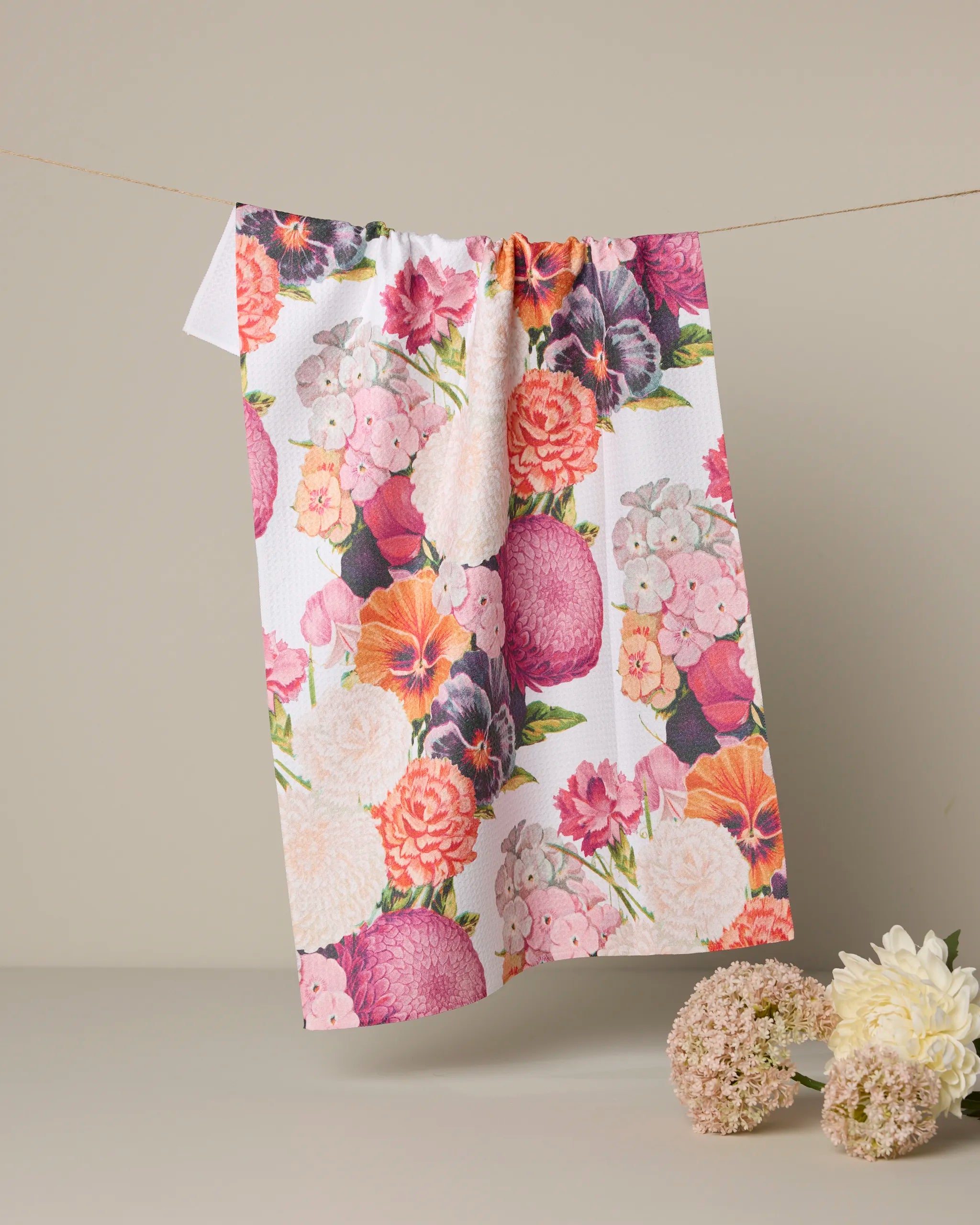 The Geometry "Ecstatic Blooms" tea towel, crafted from recycled materials, displays a vibrant pink, purple, orange, and cream floral pattern. Shown hanging against a neutral background with fresh flowers nearby.