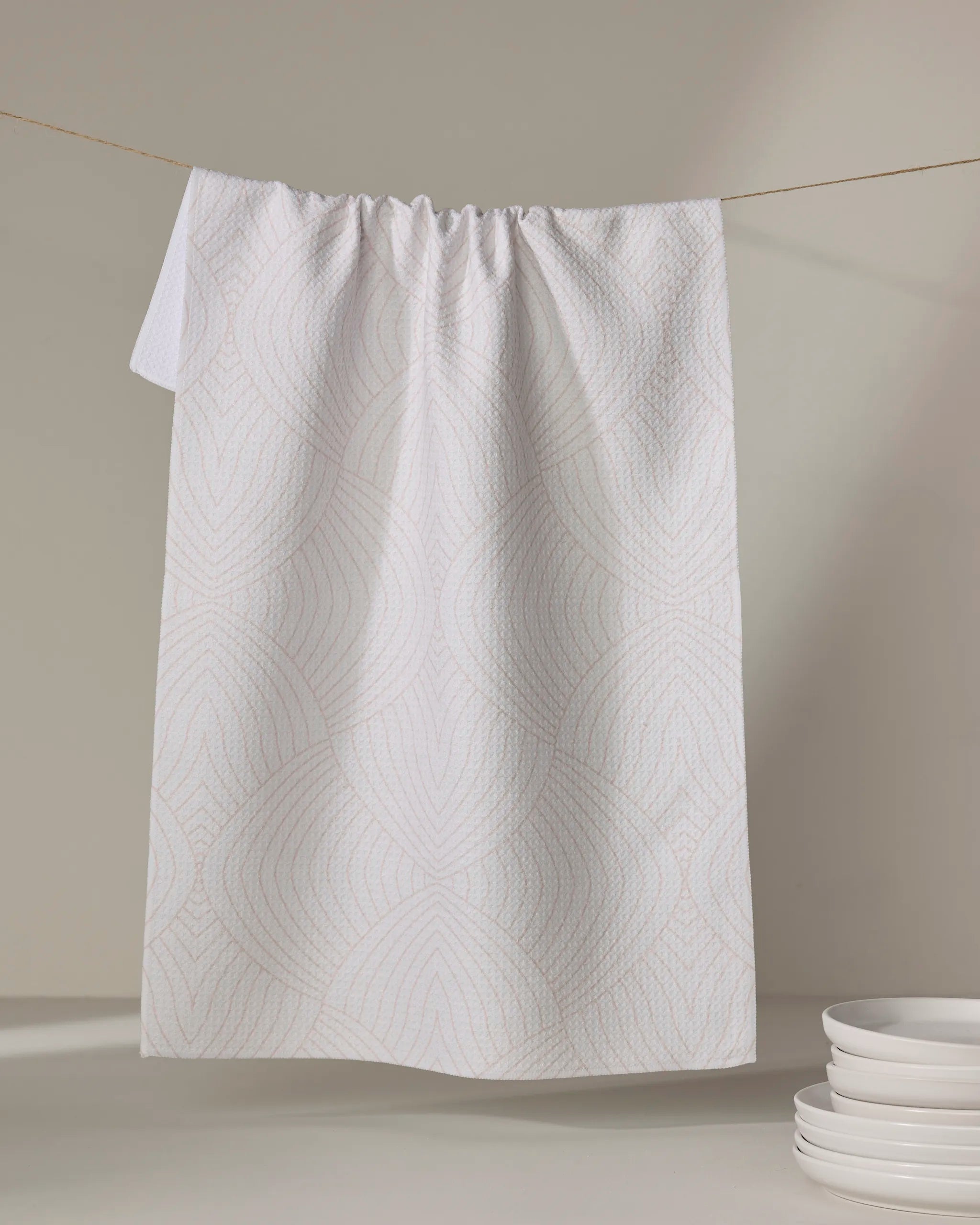 The Eileen tea towel by Amy MacCready, made from recycled materials with a white textured wavy geometric pattern, hangs from a line next to a stack of white plates on a light surface.