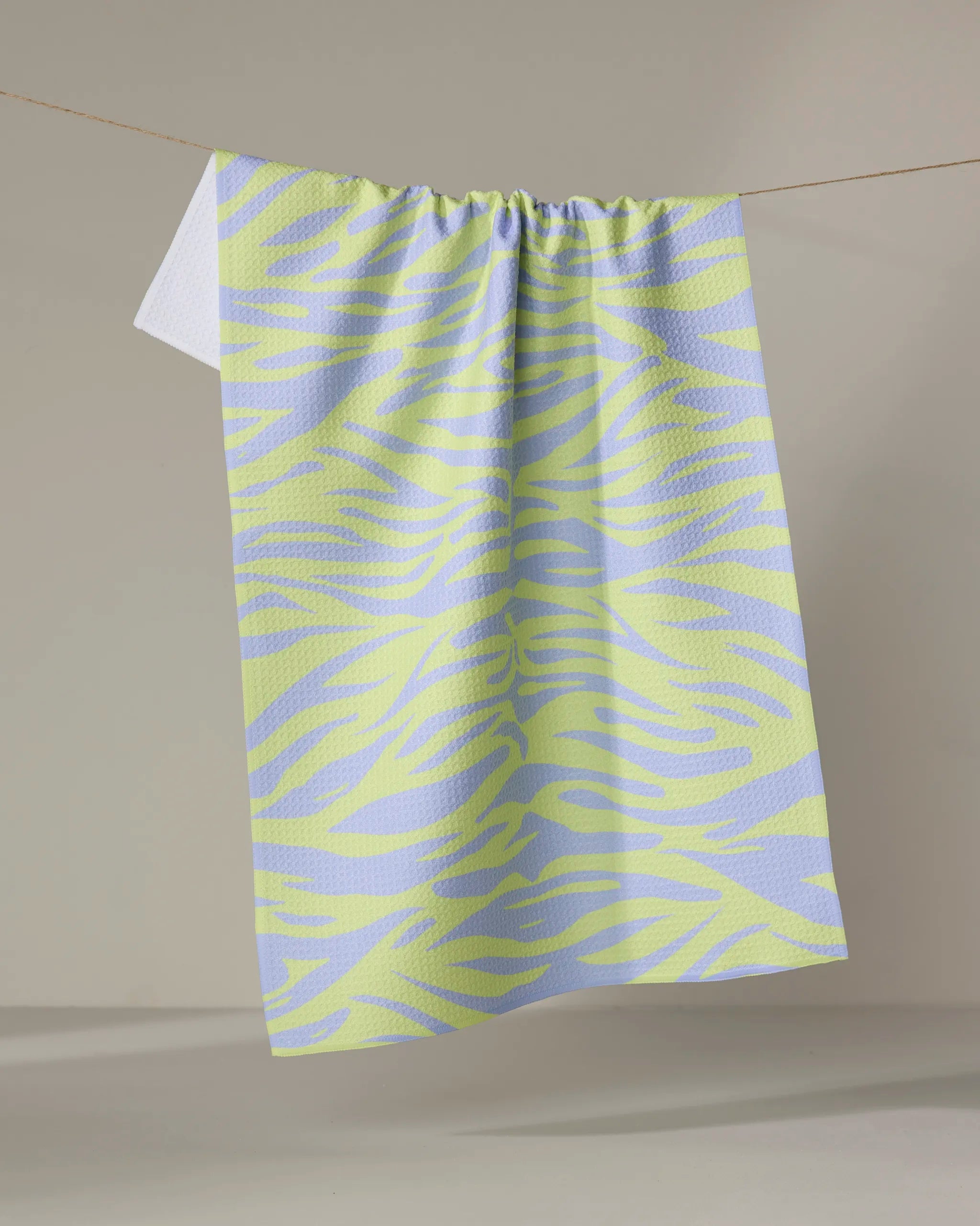 The Electric Tiger tea towel by Elisabeth Wing, featuring light purple and yellow zebra stripes and made from recycled materials, hangs on a clothesline against a neutral backdrop.