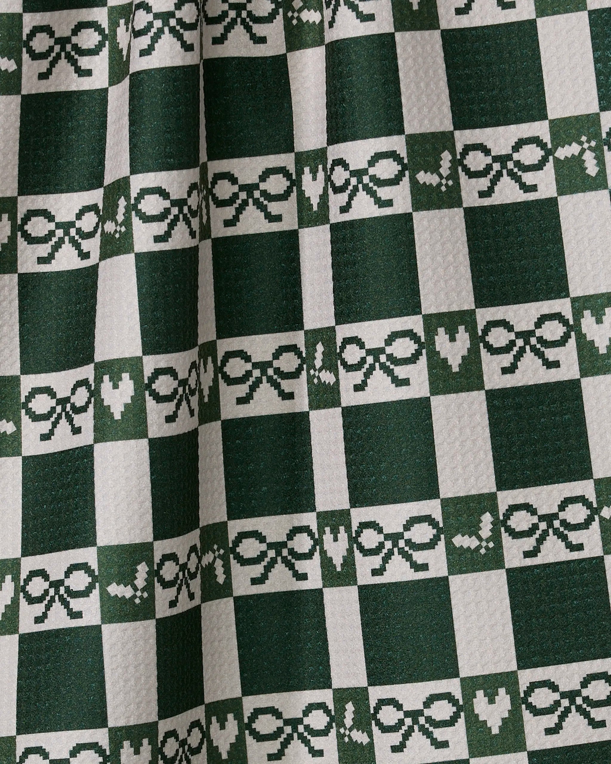 A close-up of the Geometry Elf Gingham tea towel, ultra-absorbent and made from recycled materials, shows its green and white checkered pattern with pixelated bows and hearts and slight fabric folds.