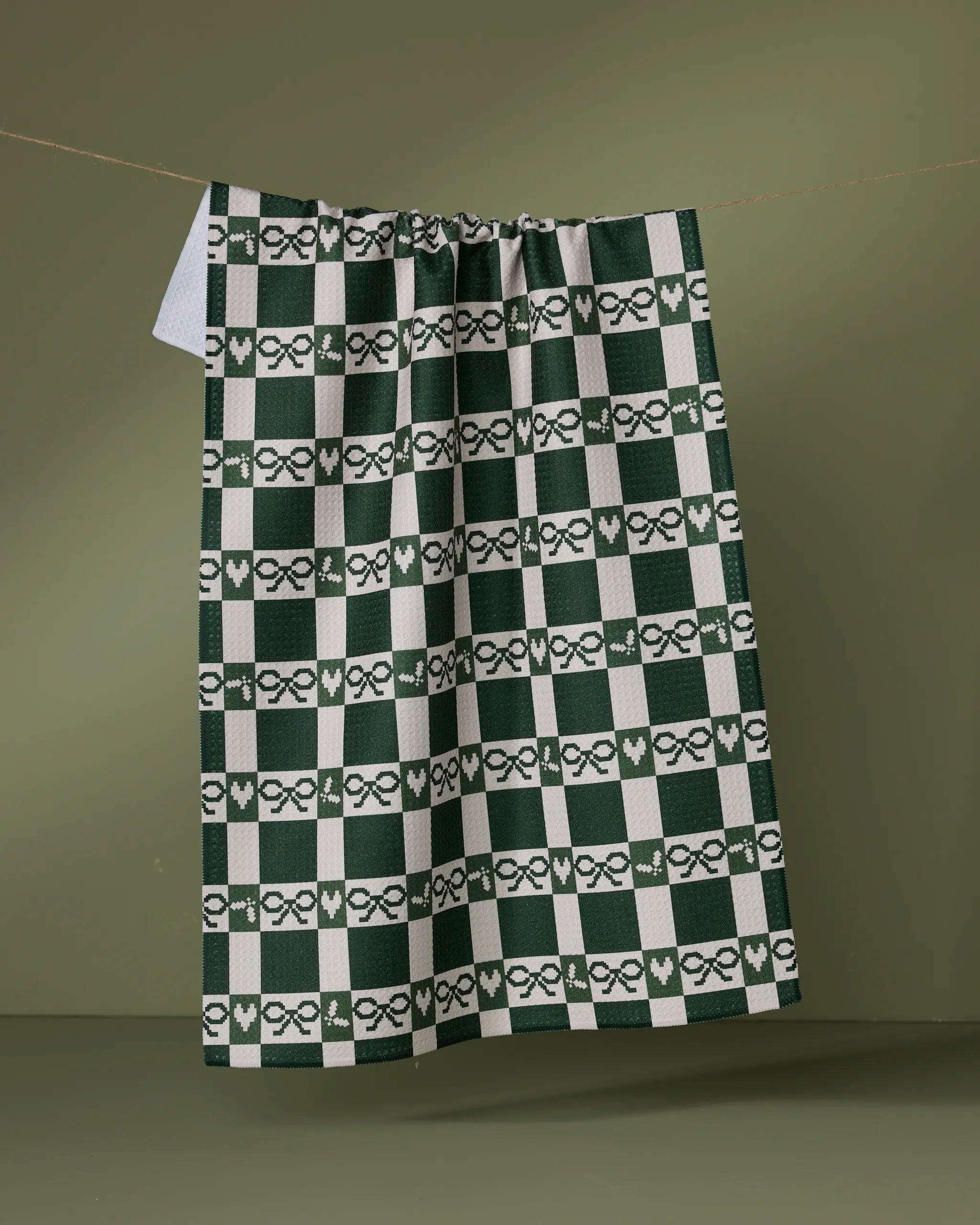 The Elf Gingham tea towel by Geometry features green and white checks with bow and heart designs on an olive background, made from recycled materials and perfect for adding charm to your kitchen.
