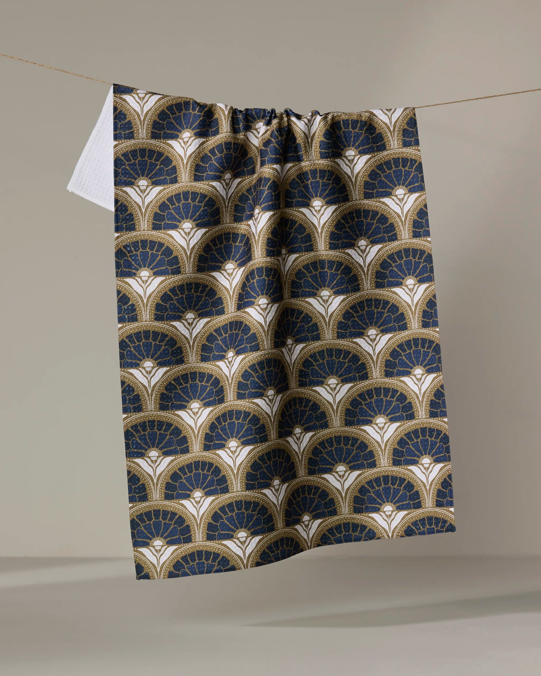The Eliza tea towel by Amy MacCready, featuring a quick-drying art deco fan pattern in navy blue, gold, and white, hangs on a string against a neutral background.