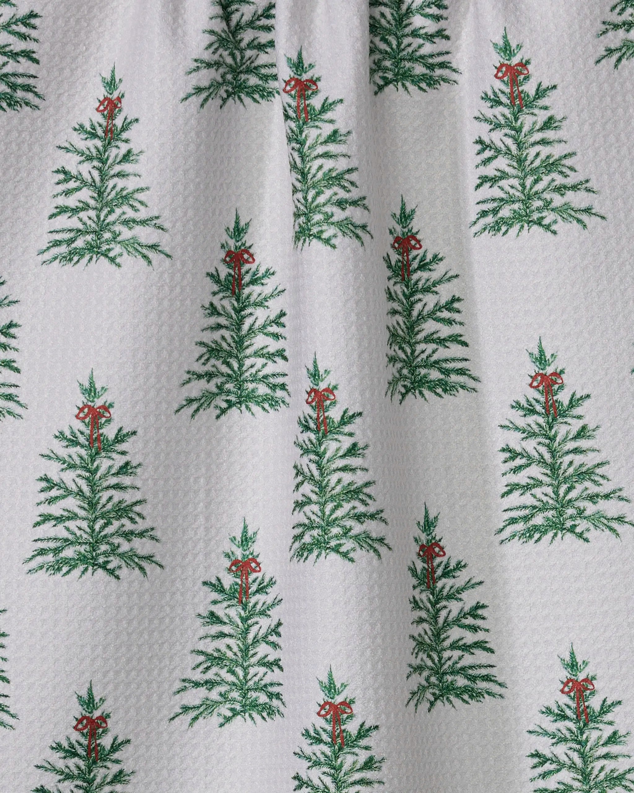 The Geometry Ellsworth tea towel features a white fabric with evenly spaced green Christmas trees topped with red bows. Made from recycled materials, it offers a festive and eco-friendly touch for your holiday decor.