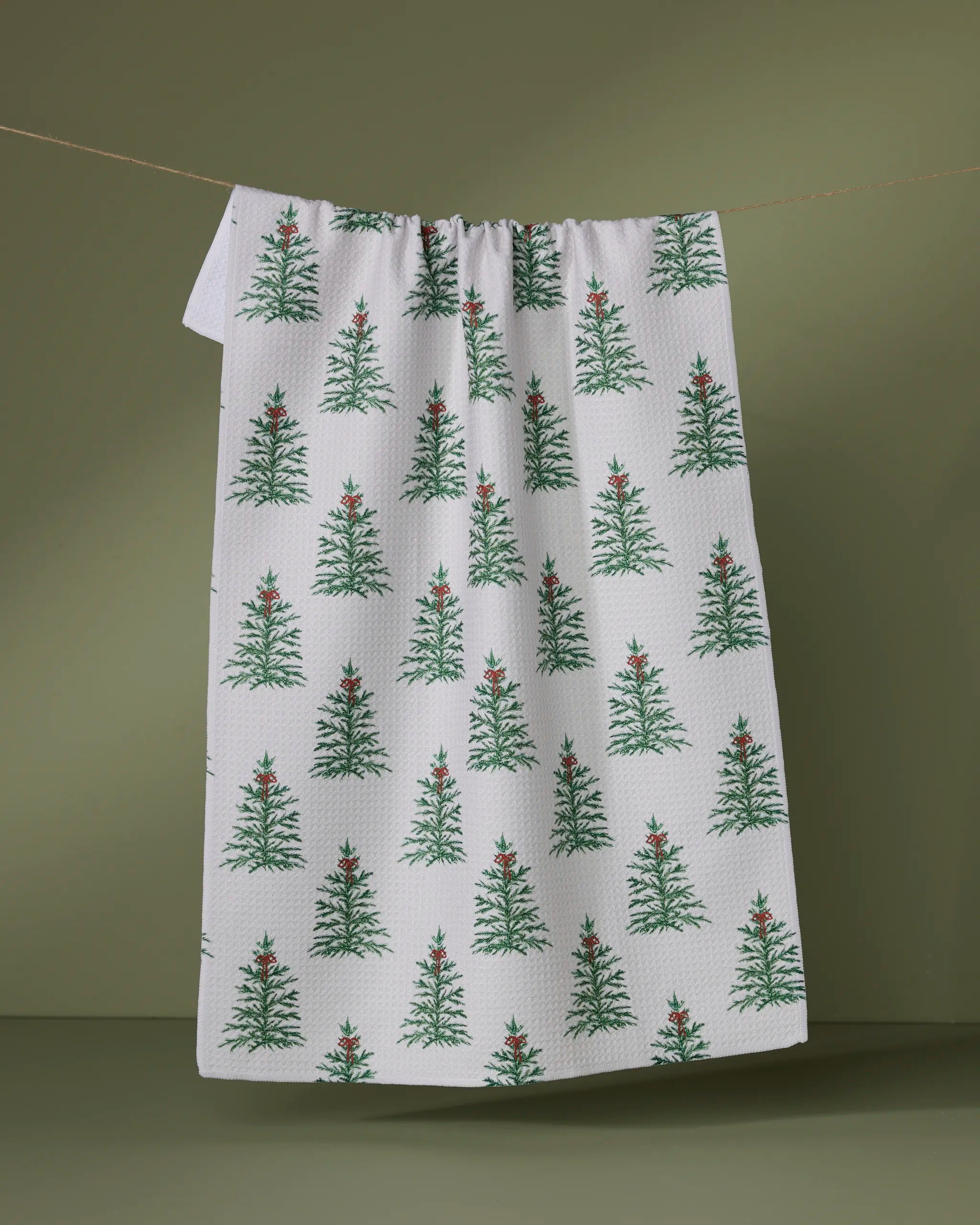 The Geometry Ellsworth tea towel, made from recycled materials, displays green Christmas trees with red stars and hangs on a clothesline set against an olive-green background.
