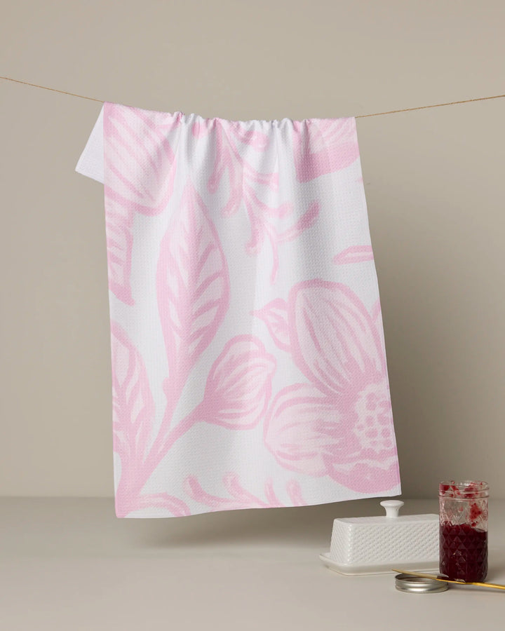 The Elsie tea towel by Geometry, featuring a large pink botanical print on white and made from recycled materials, hangs on a line against a beige background. Below are a butter dish, plate with knife, and jar of red jam.