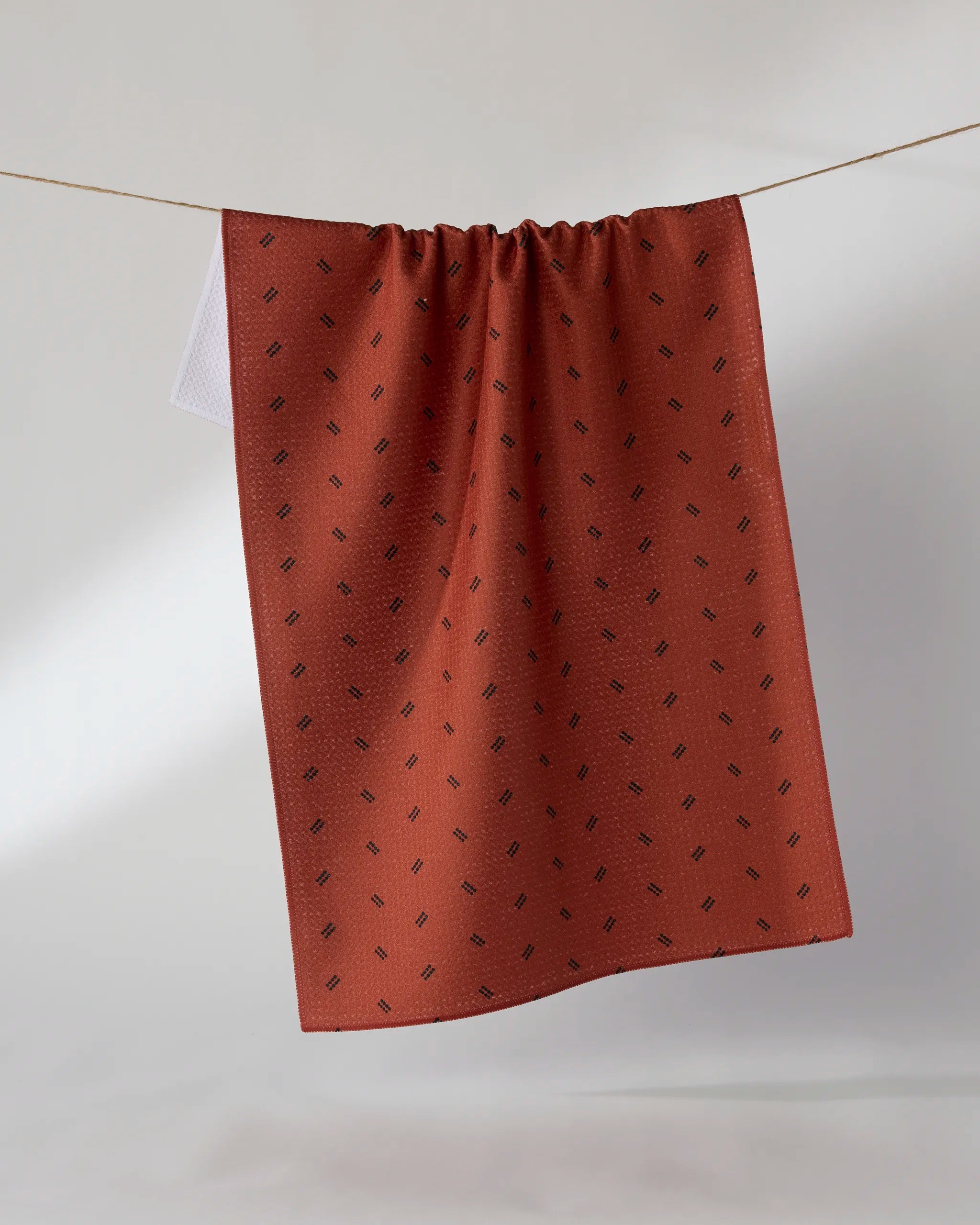 A Geometry Ember Dot tea towel in rust-colored Geoweave fabric, featuring a small repeating geometric pattern, hangs over a clothesline against a plain light background.