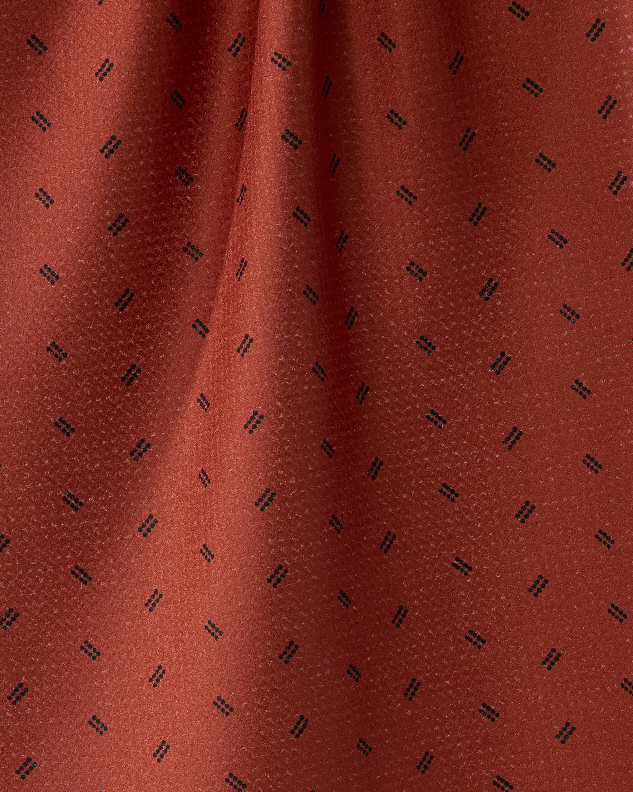 A close-up of Geometry's Ember Dot Geoweave fabric shows rust tones with a subtle sheen and pairs of small black diagonal lines, likely from a recycled material tea towel, appearing slightly gathered or draped.