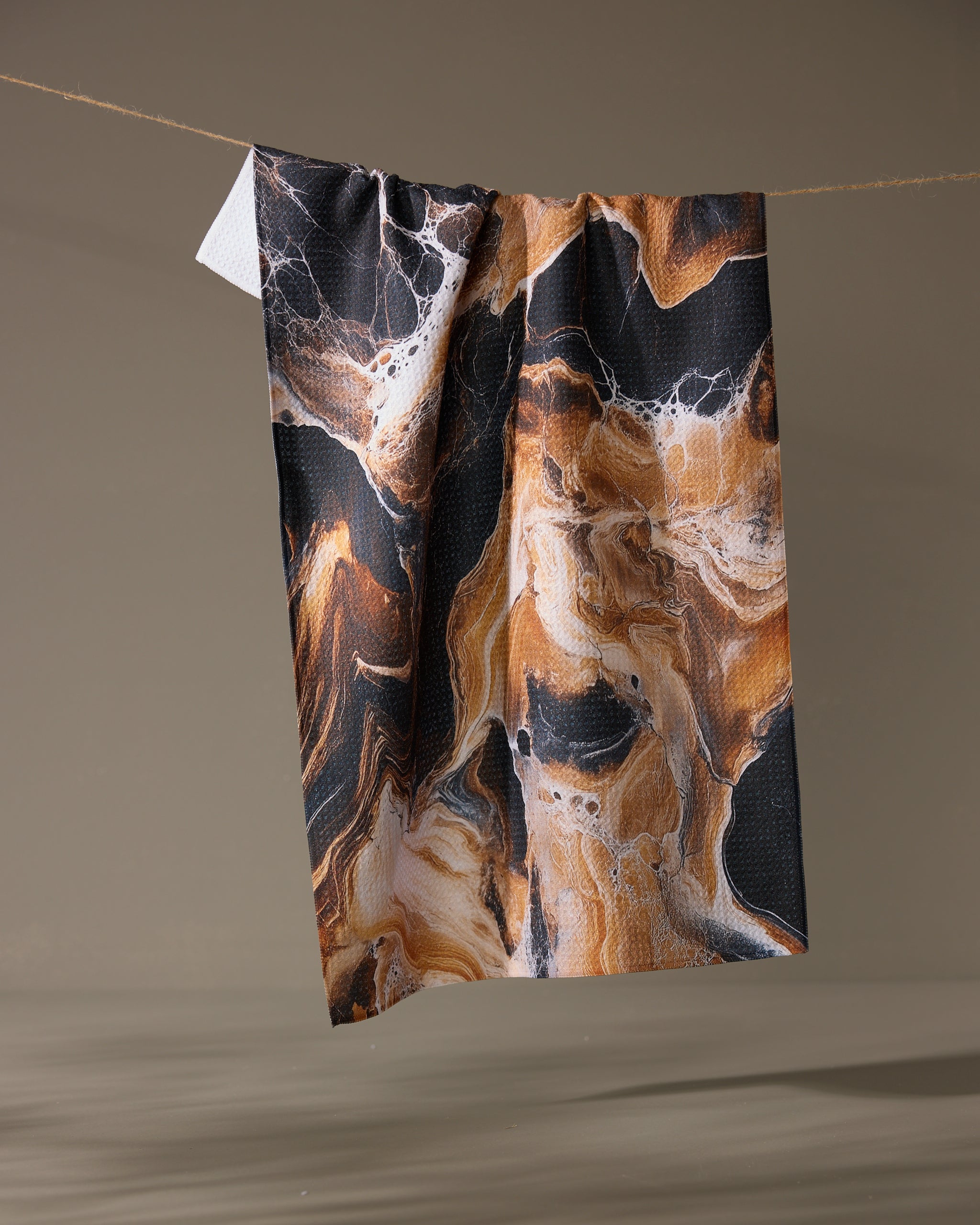 The Geometry Emperador recycled tea towel, featuring a black, brown, and white marbled design, hangs from a string against a plain beige background.