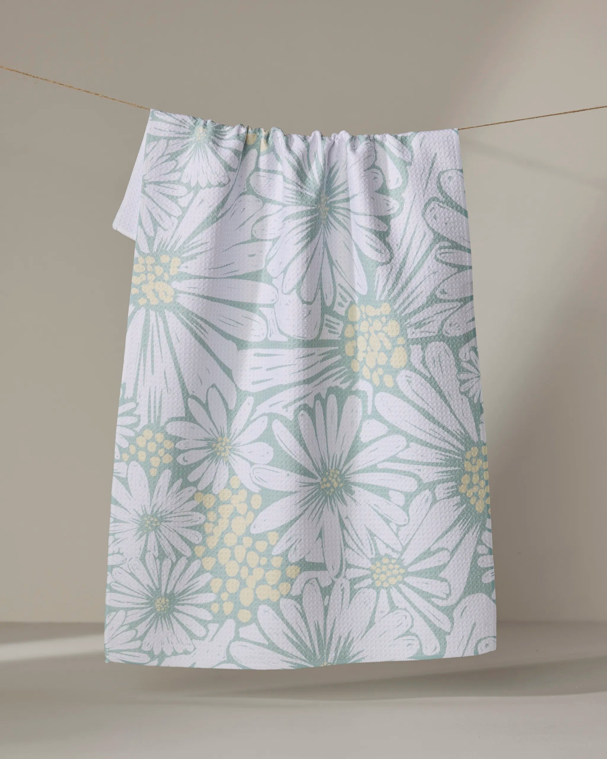 The English Daisies tea towel by Elisabeth Wing, featuring a quick-drying pastel floral design with large white and yellow flowers, hangs from a clothesline against a neutral background.