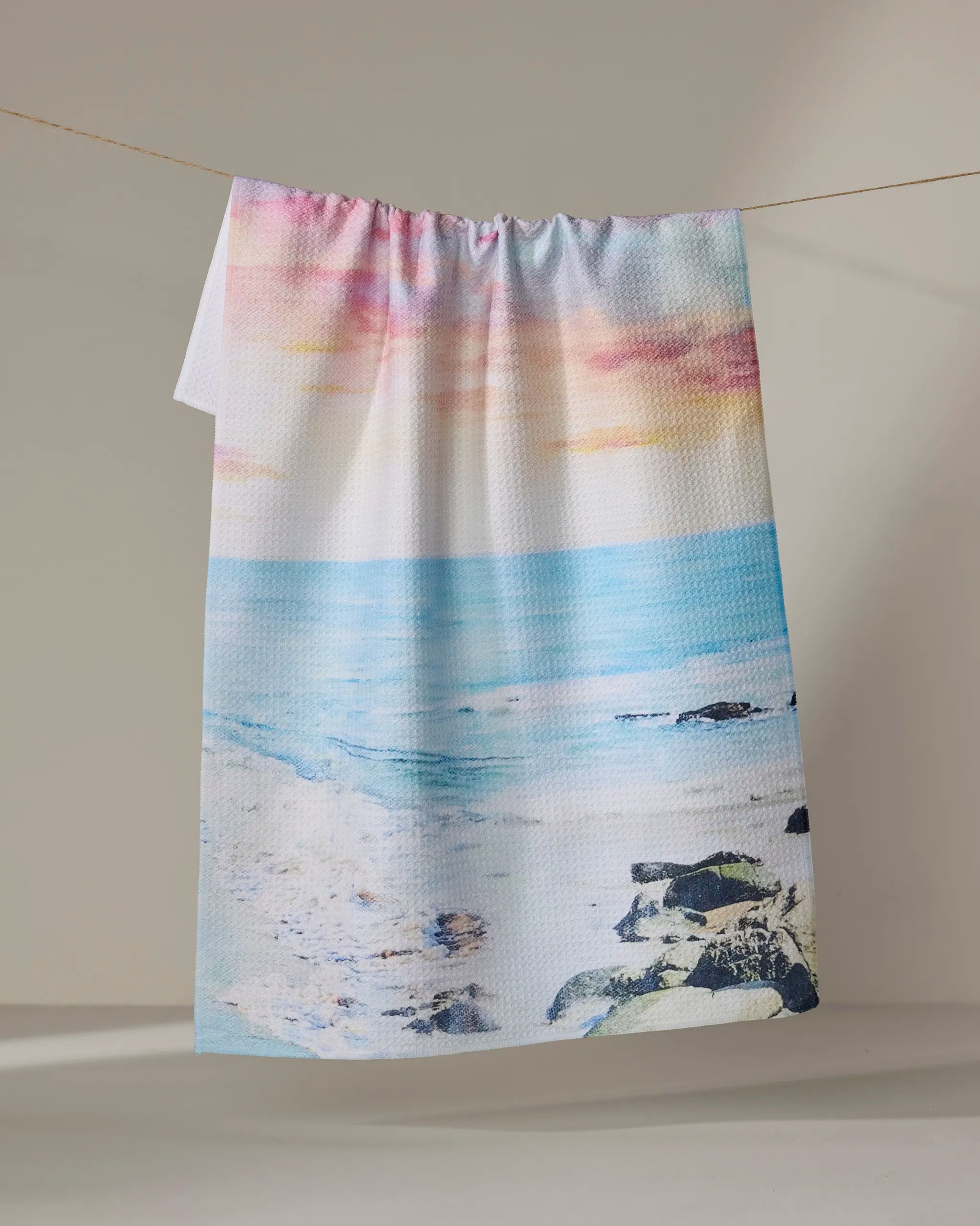The Stefanie Bales "Epiphany" towel, featuring a beach scene with blue ocean waves, rocks, and a pastel sky, hangs on a clothesline against a neutral background.