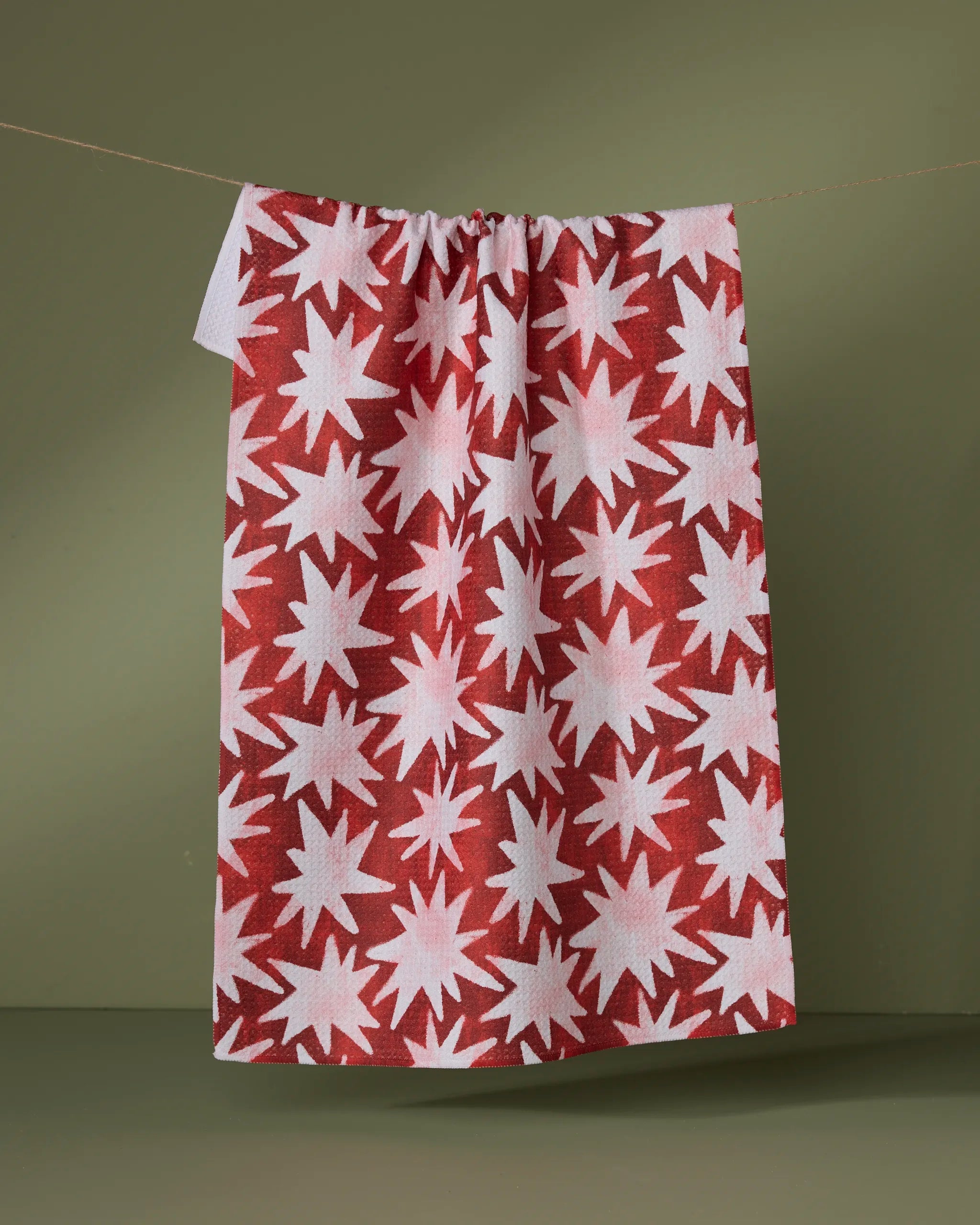 The Estrellas tea towel by Geometry, featuring bold red and white starburst patterns, hangs from a string against a plain olive-green background.