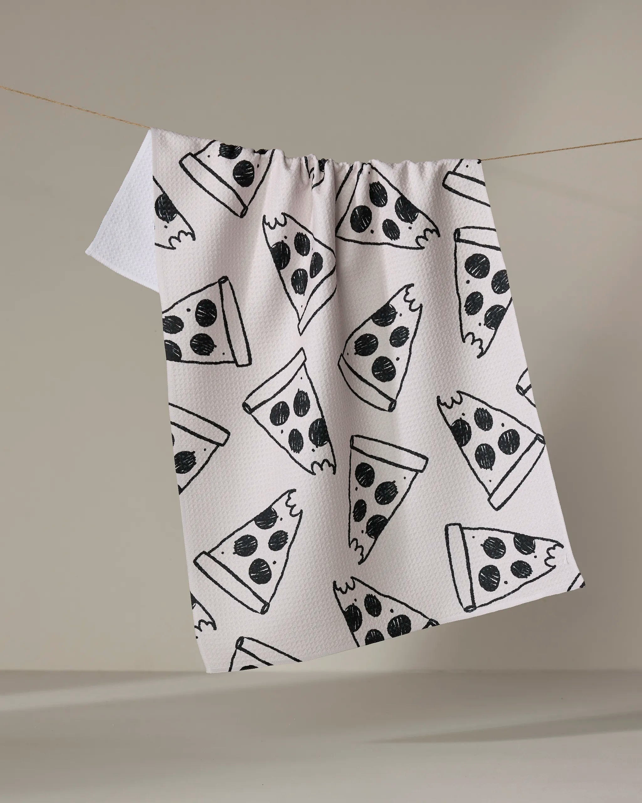 The Cole Strem Eternal Slice towel, featuring a black pepperoni pizza slice pattern on ultra-absorbent white fabric, hangs on a clothesline against a plain, neutral background.