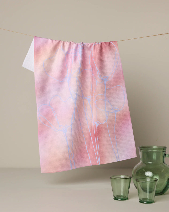 Geometry's Ethereal Bloom tea towel in pink and peach, featuring a subtle abstract floral design, hangs above two small green recycled glass cups and a matching vase—a sustainable, elegant addition to your kitchen.
