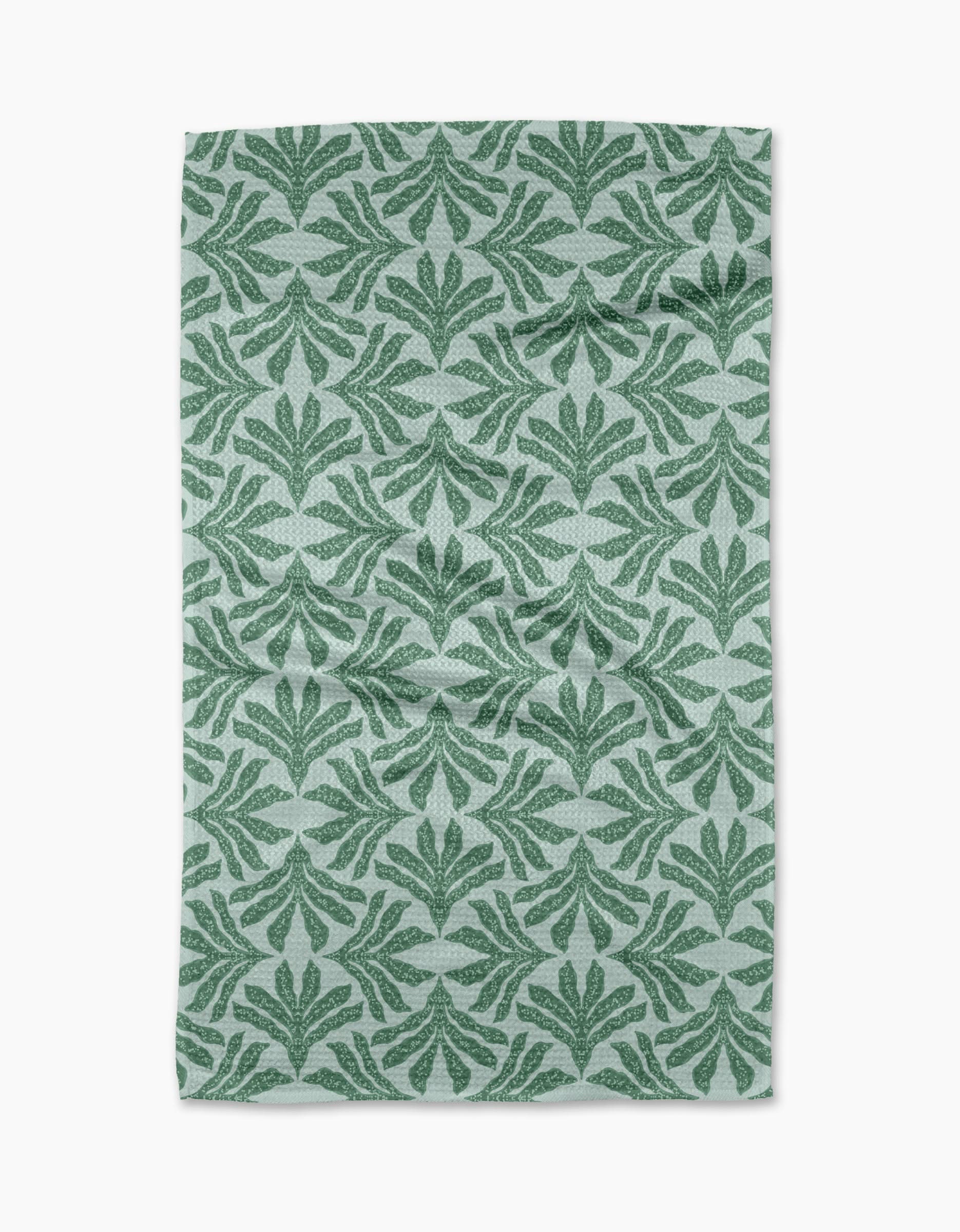 The Ethereal Seaweeds tea towel by Jasmine Blooms Designs features a green leafy pattern on light gray recycled fabric with symmetrical stylized leaf motifs. The slightly wrinkled towel is displayed against a white background.