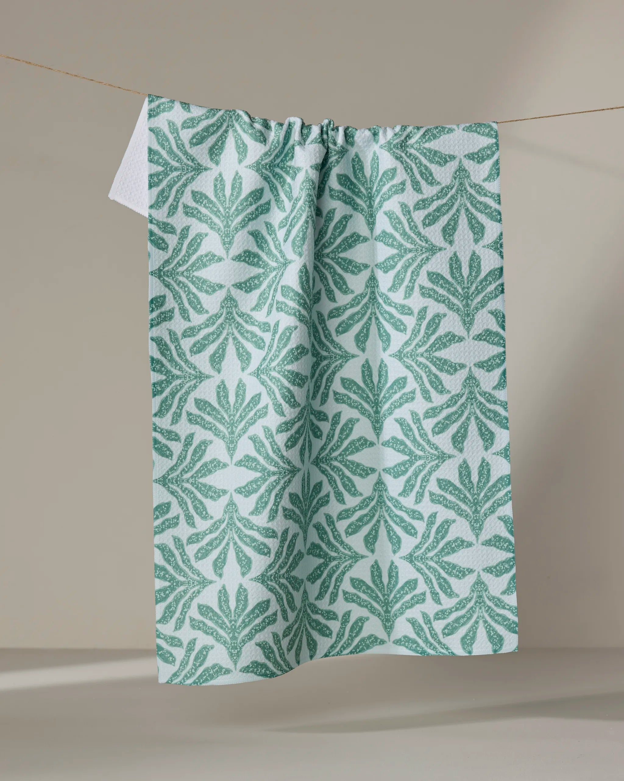 The Ethereal Seaweeds tea towel by Jasmine Blooms Designs, featuring a green leafy pattern on light blue fabric, hangs on a clothesline against a plain beige background.