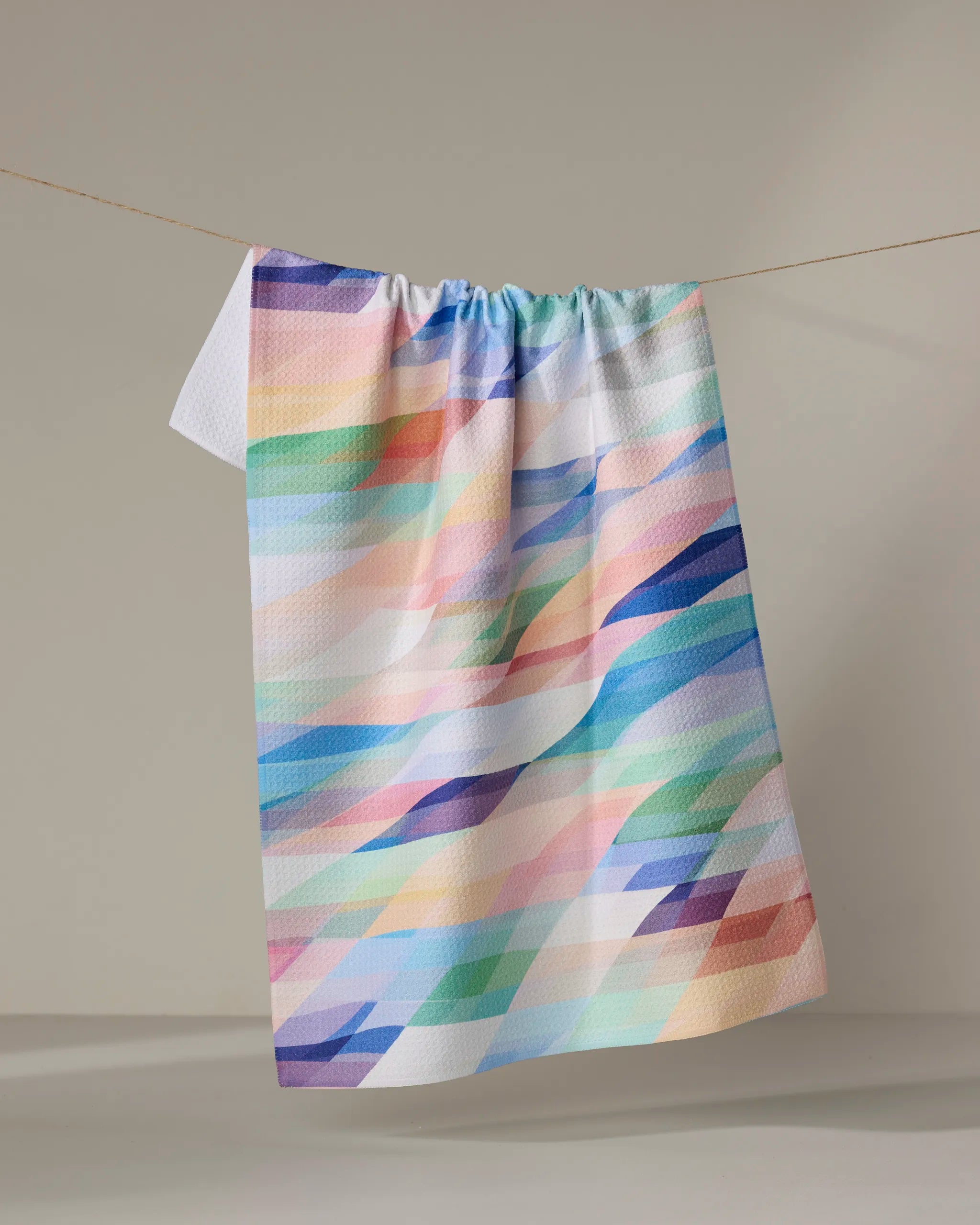 The Eventide tea towel by Rachel Parker, featuring a pastel geometric pattern and quick-drying fabric, hangs on a clothesline against a neutral background.