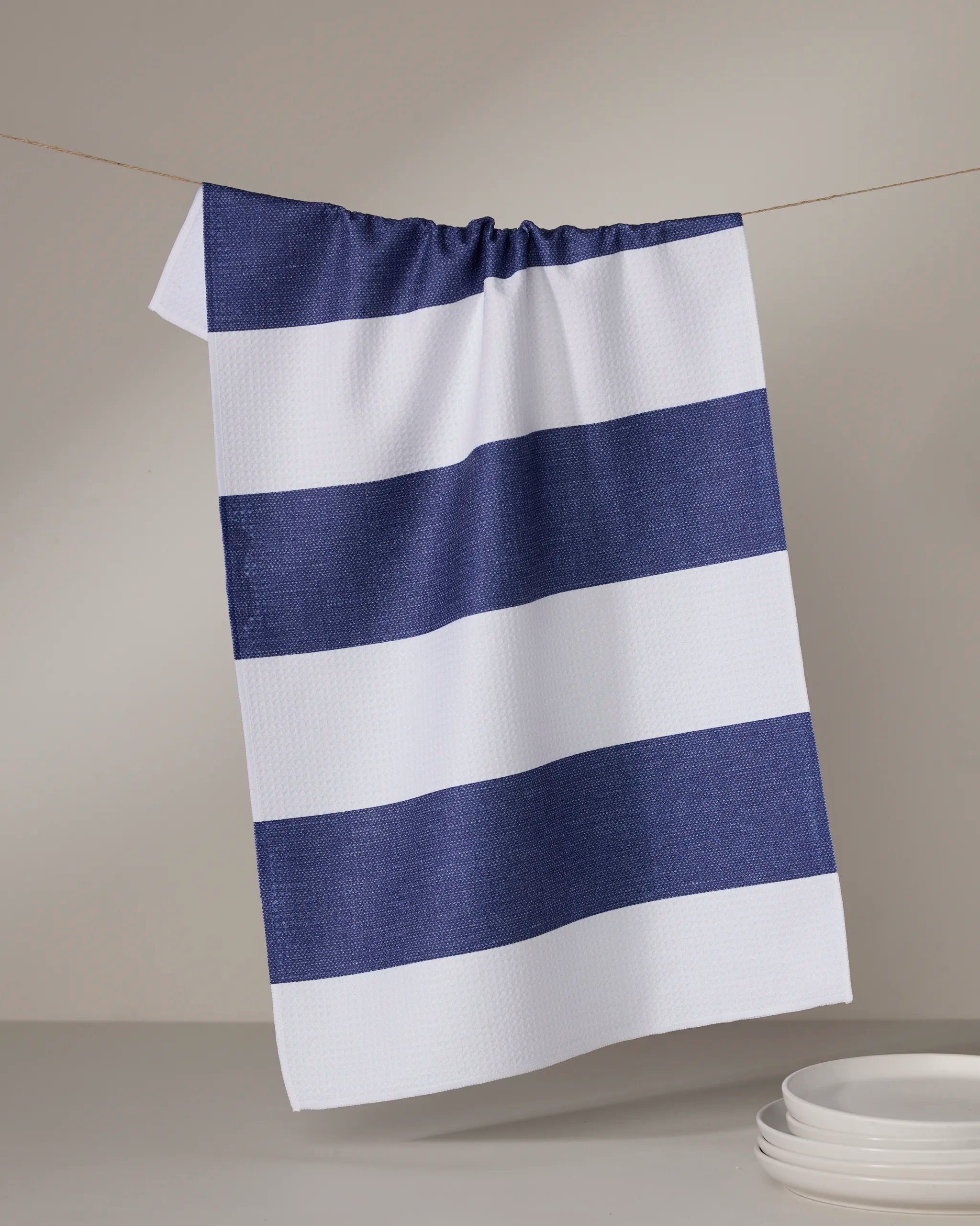 A Geometry Everband kitchen towel, made from recycled materials, in white and navy blue stripes hangs on a string above stacked white plates with a plain, light background.