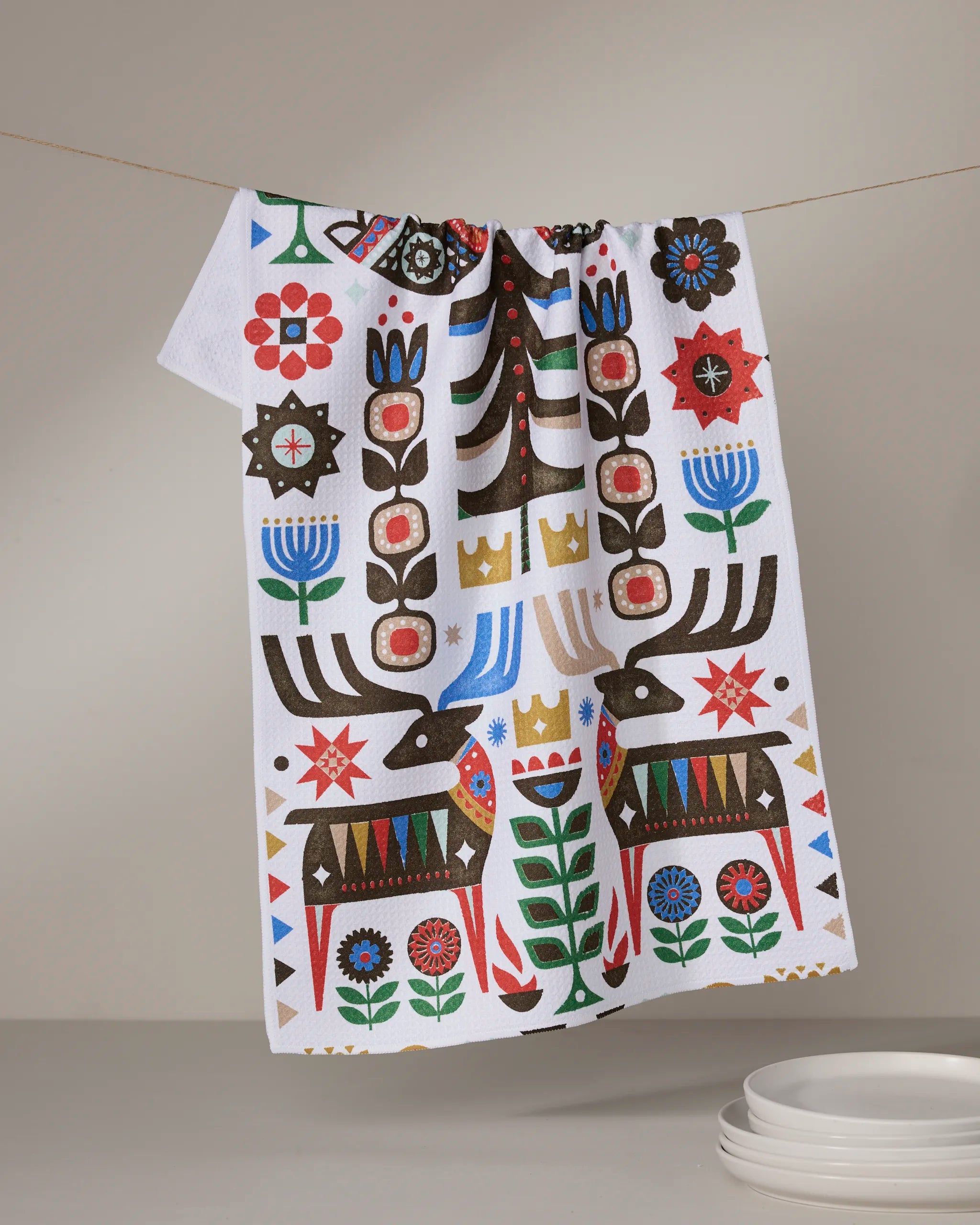 The Geometry Everfolk tea towel, ultra-absorbent and white with a vibrant folk art pattern of stylized deer, plants, crowns, and geometric shapes in red, blue, green, and brown, hangs above stacked white plates.