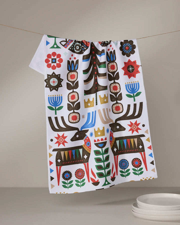 The Geometry Everfolk tea towel, ultra-absorbent and white with a vibrant folk art pattern of stylized deer, plants, crowns, and geometric shapes in red, blue, green, and brown, hangs above stacked white plates.