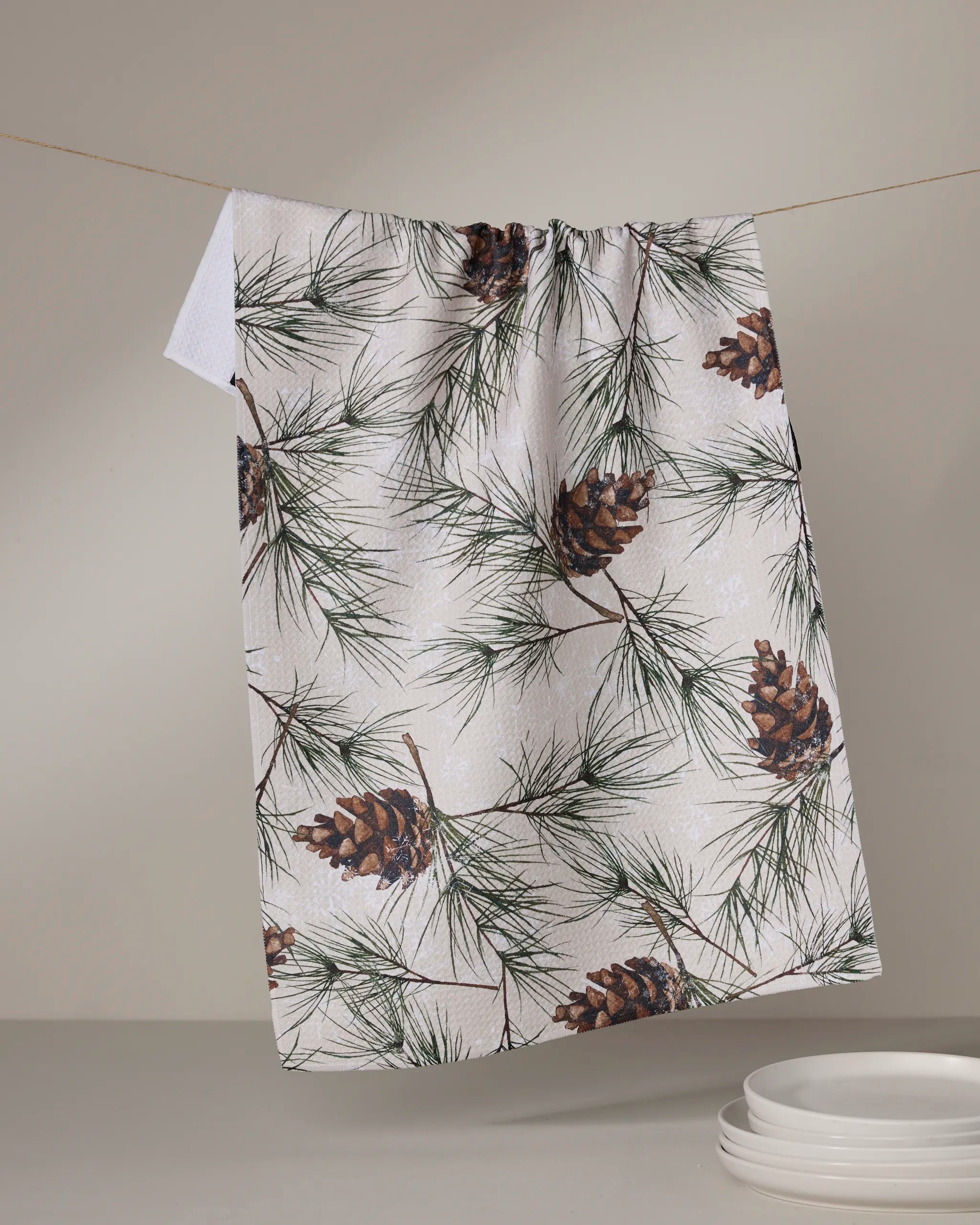 The Geometry "Evergreen Frost" kitchen towel, featuring a pinecone and pine needle pattern, hangs from a clothesline above a stack of white dishes on a light-colored surface.