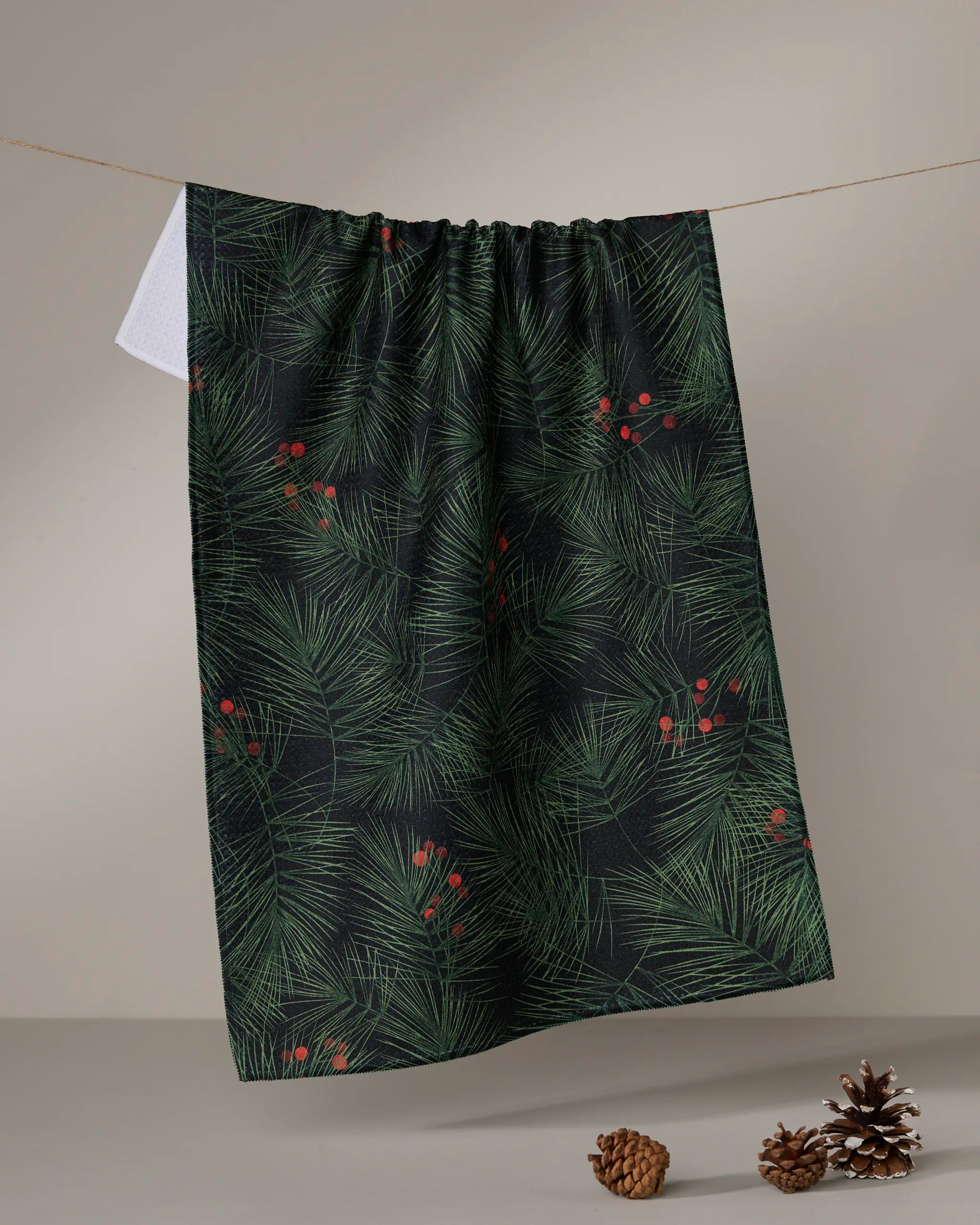 The Geometry Evergreen Pine ultra-absorbent tea towel, featuring a dark background with green pine needles and red berries, hangs on a string above three pinecones against a plain beige backdrop.