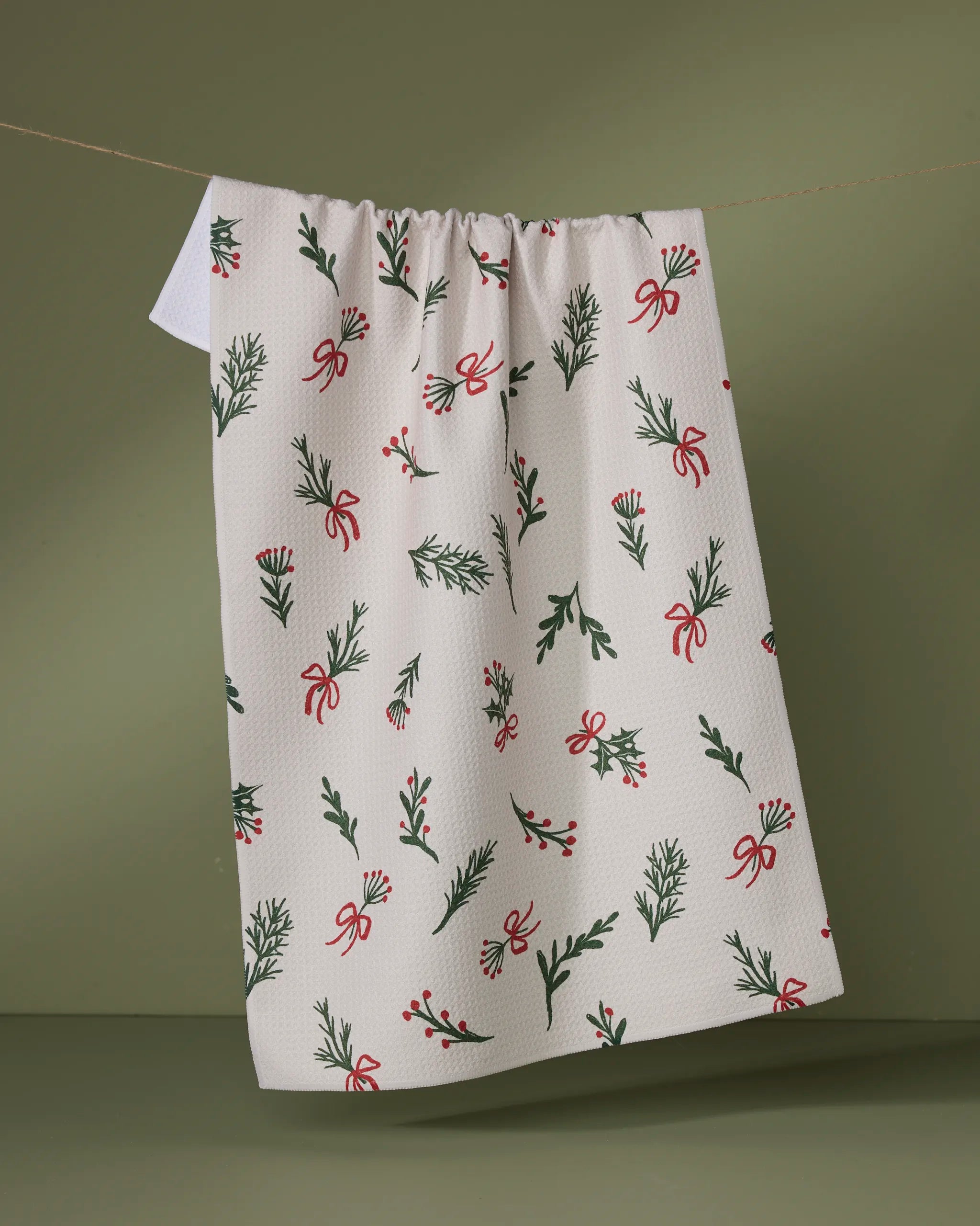 The Geometry Everlove kitchen towel features a festive pattern of green pine branches, red berries, and red bows, and hangs on a clothesline against an olive green background.