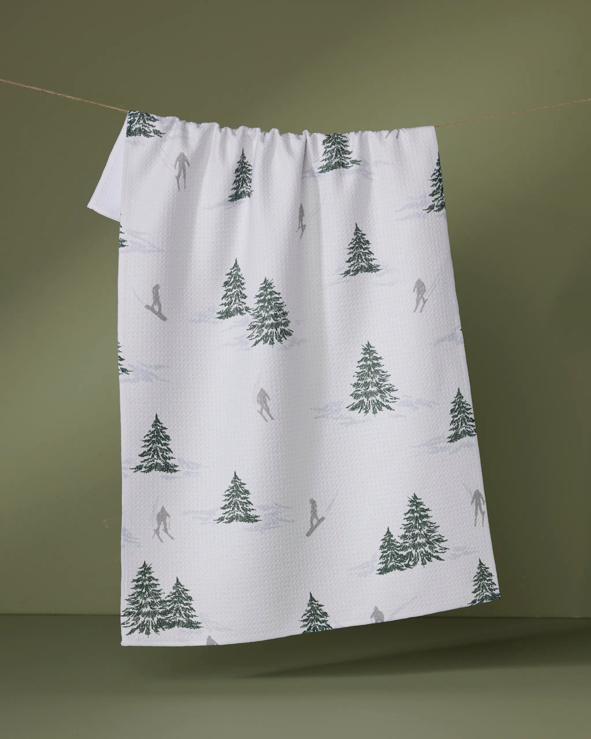 The Geometry Evermont towel, featuring green pine trees and gray skier silhouettes, hangs on a line against an olive green background.