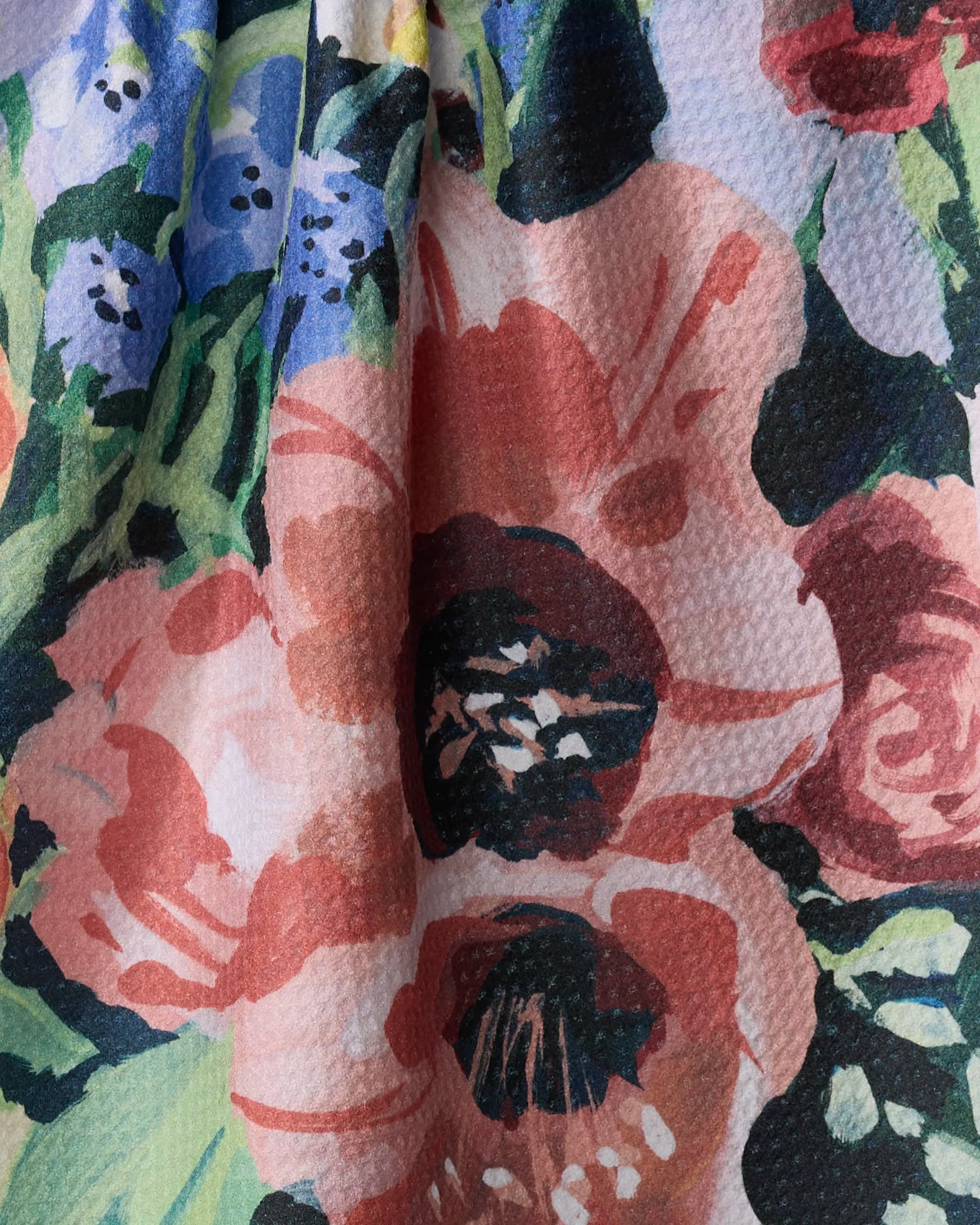 Close-up of Exotic Bouquet tea towel fabric by Ania Zwara Art, featuring a colorful floral pattern—large red, pink, and purple flowers with green leaves plus black, white, and blue accents. The top edge of the textured fabric appears slightly gathered.