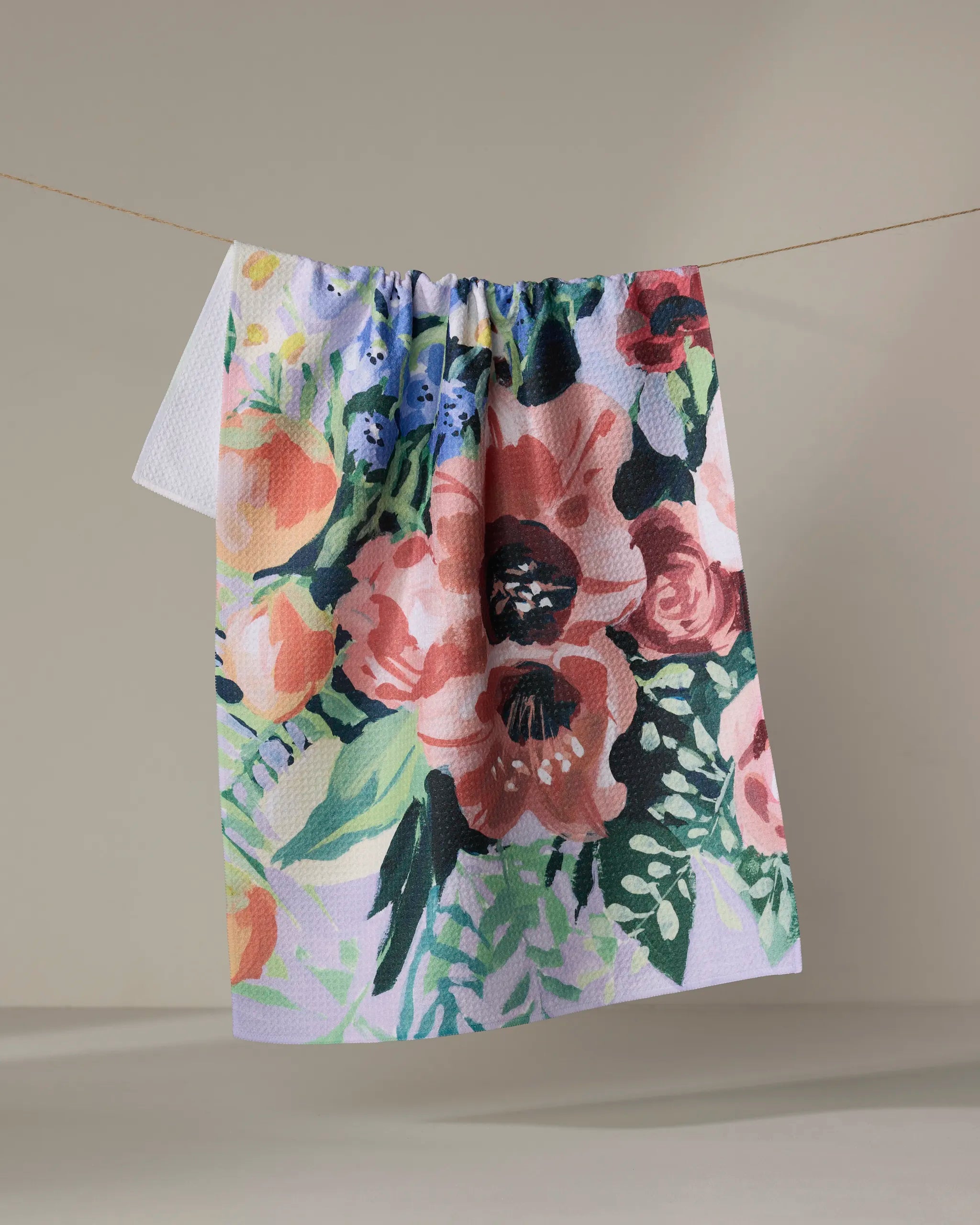 The Exotic Bouquet tea towel by Ania Zwara Art, made from recycled materials, features a vibrant floral pattern—pink, red, purple, and blue flowers with green leaves—hanging on a clothesline against a neutral backdrop.