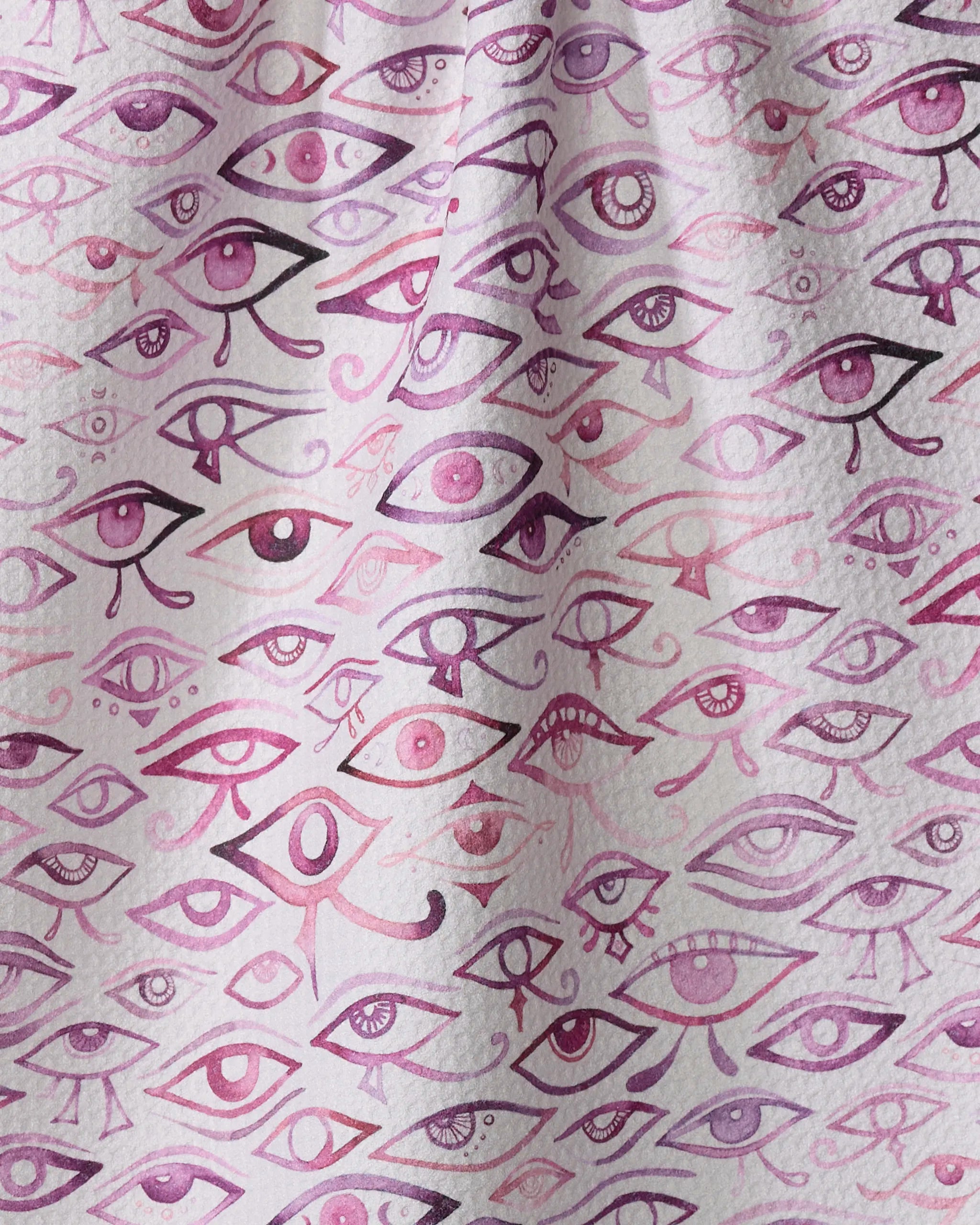 Eye Of Horus by Maayan Alper-Swan features a quick-drying fabric densely covered in purple and pink illustrated eyes of various shapes and sizes, creating a vibrant tea towel effect with visible folds in the material.