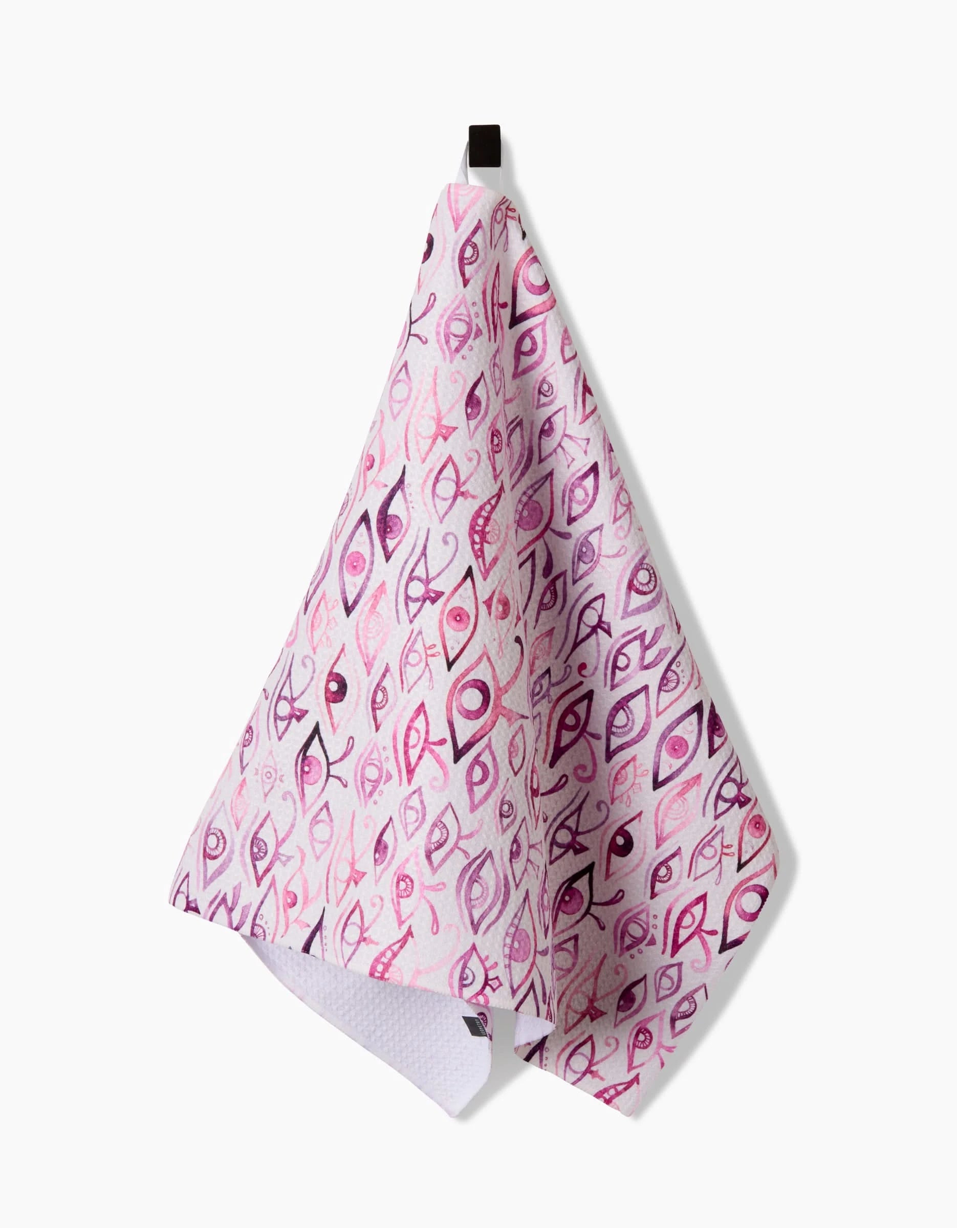 The Eye Of Horus tea towel by Maayan Alper-Swan, featuring abstract fish designs in pink and purple Geoweave fabric, hangs from a small black hook against a plain white background.