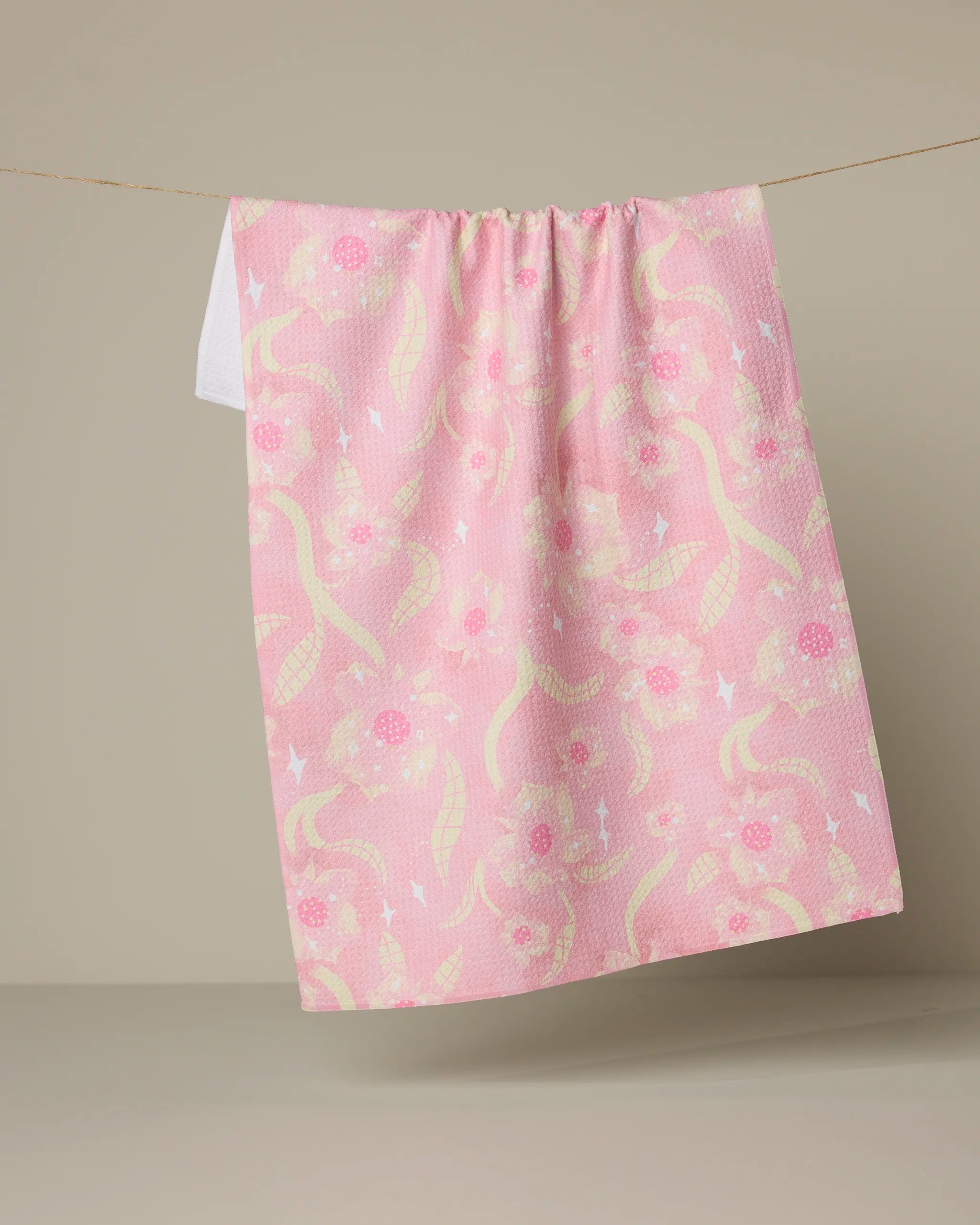 The Geometry Fairy Dust Floral tea towel, featuring a pink and white floral and leaf pattern, hangs from a string against a neutral beige background.