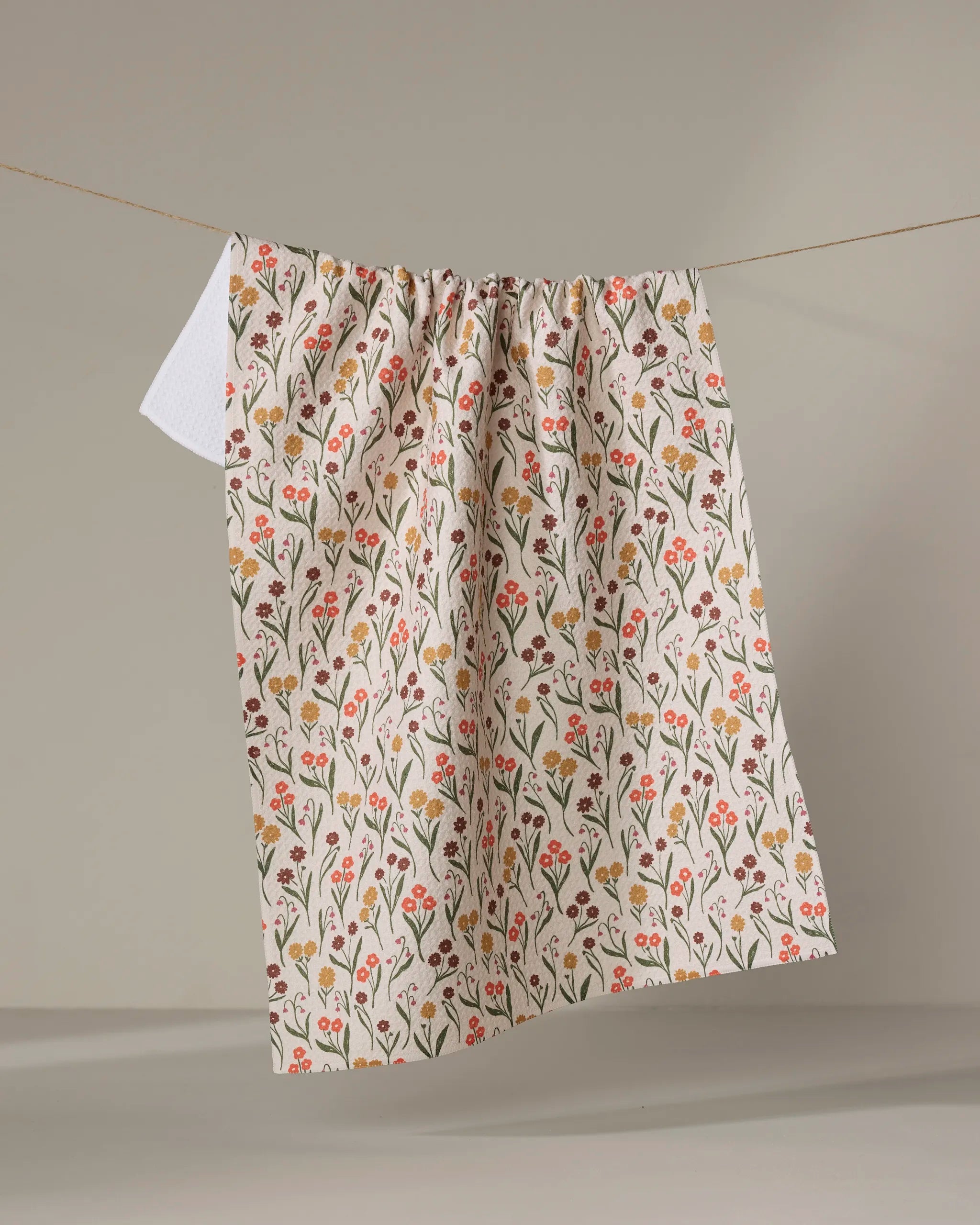 The Fall Blossom kitchen towel by Kayla Ann, featuring red, yellow, and orange floral patterns and made from recycled materials, hangs on a clothesline against a neutral background.