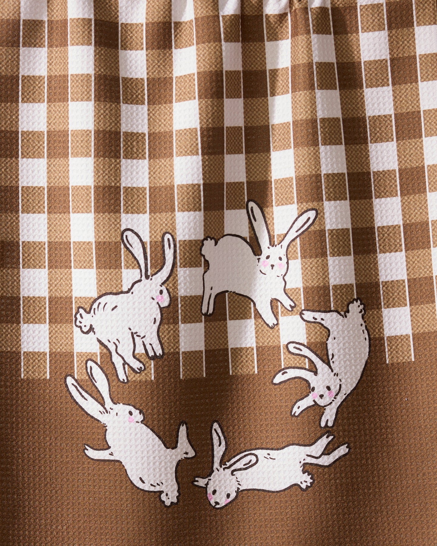 The Geometry Fall Bunnies tea towel features quick-drying recycled fabric in a brown and white checkered pattern, with five white cartoon rabbits with pink cheeks playfully circling at the bottom.