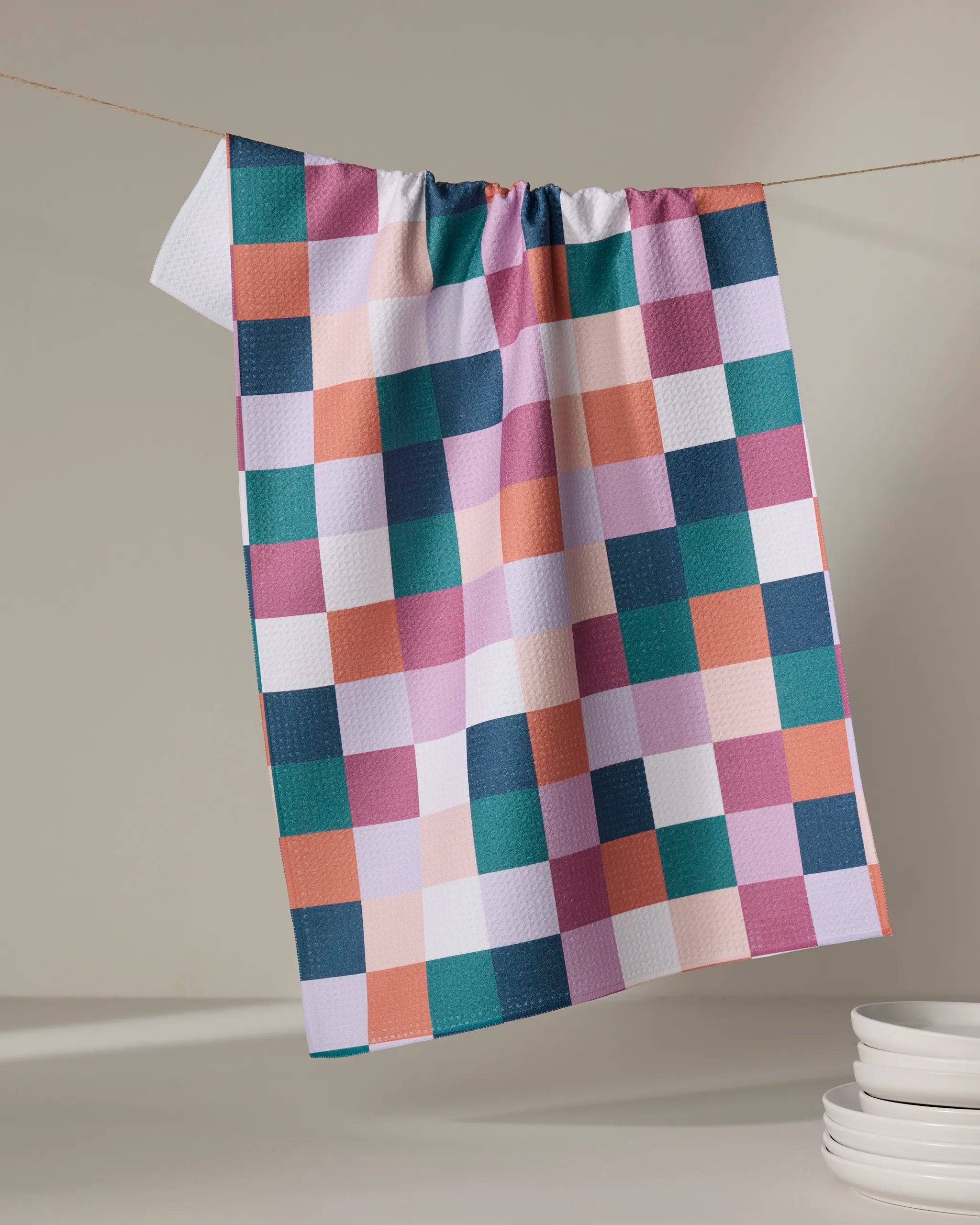 The Fall Checkers kitchen towel by Claire Iglesias, featuring pink, green, orange, navy, and white squares and made from recycled materials, hangs on a string with a stack of white plates in the lower right corner.