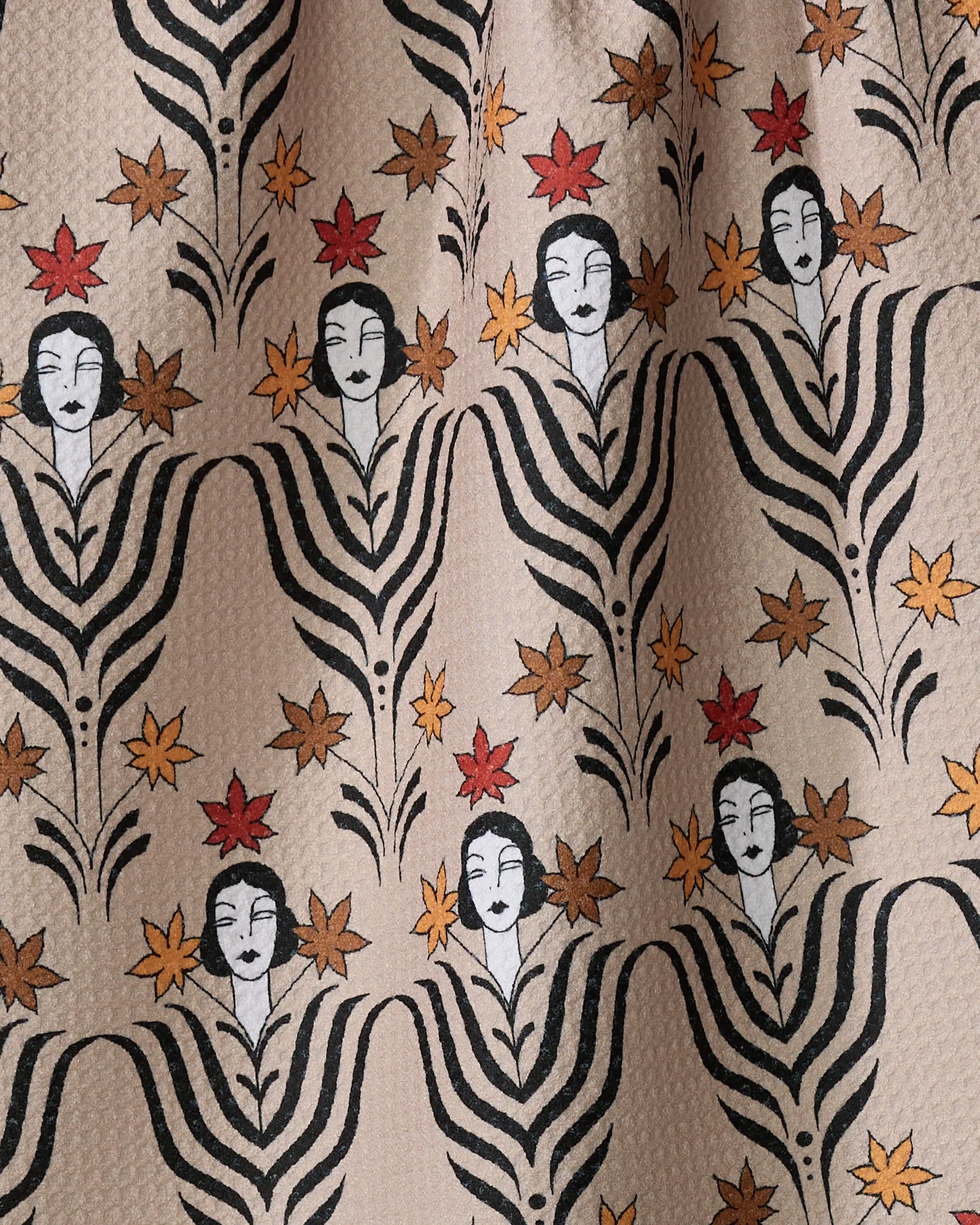 The "Fall Deco Dame" tea towel by Cole Strem features stylized black-and-white faces with flowing hair, framed by leaf-like black lines and accented with red and yellow star flowers on a beige background.