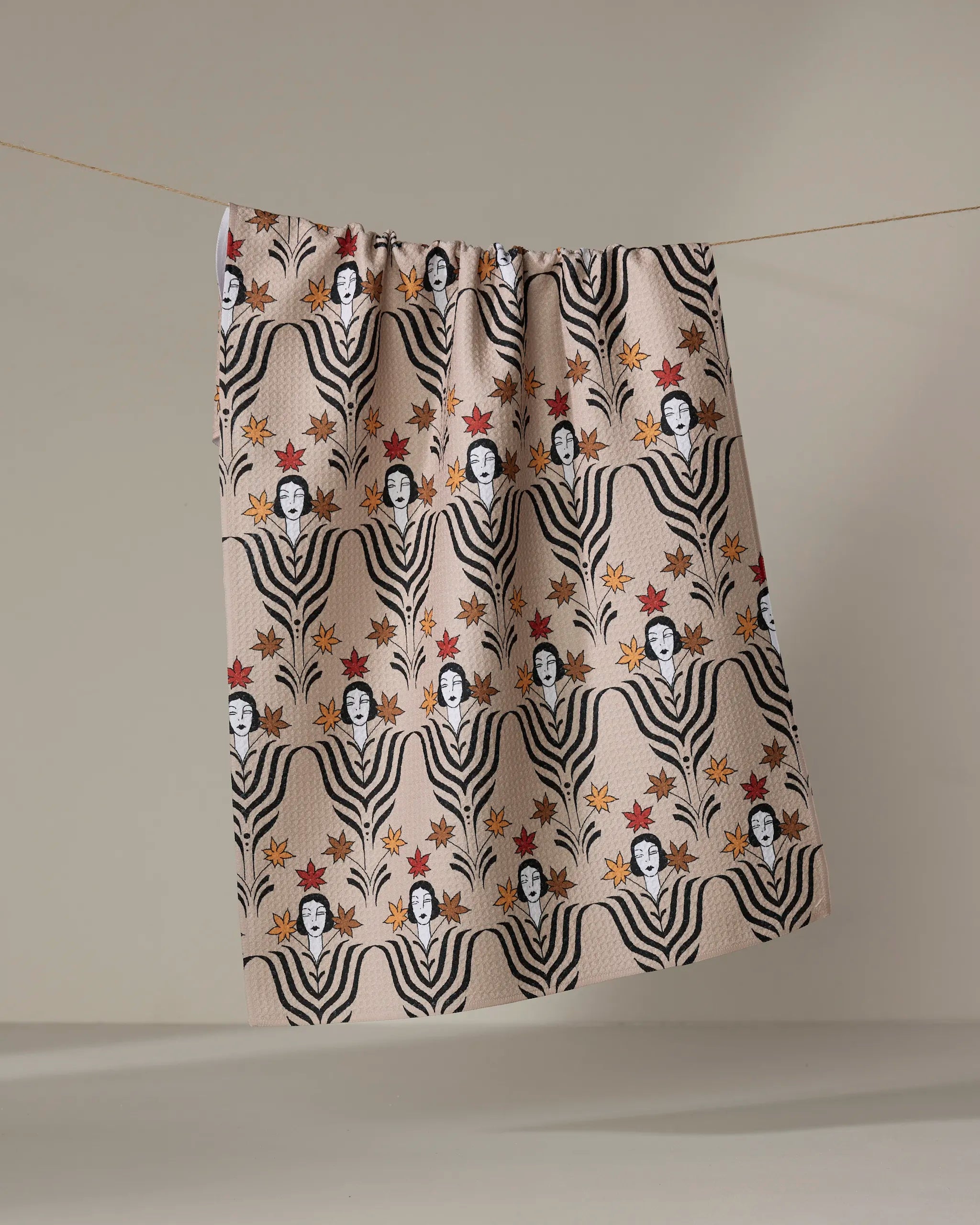 The Cole Strem “Fall Deco Dame” tea towel, made from recycled materials, features stylized black-and-white faces with wavy lines and red-orange floral accents. It hangs on a clothesline against a neutral backdrop.