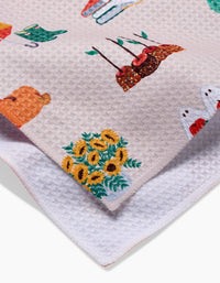 A close-up of the Fall Favorites tea towel by Ceyda Alasar, featuring textured beige fabric with embroidered sunflowers, a bear, red birds, and more. Layered over a white towel for a charming kitchen look.