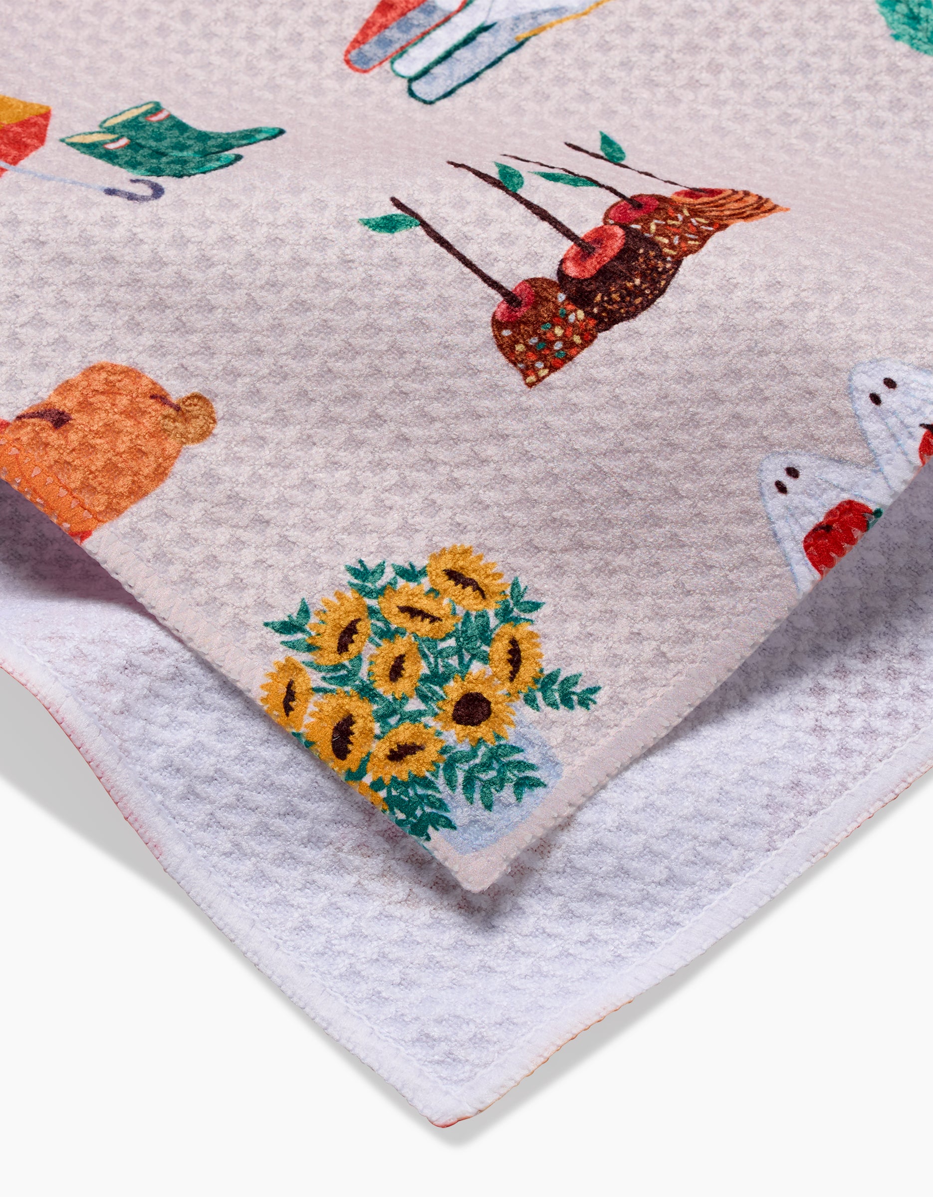 A close-up of the Fall Favorites tea towel by Ceyda Alasar, featuring textured beige fabric with embroidered sunflowers, a bear, red birds, and more. Layered over a white towel for a charming kitchen look.