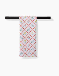 The Fall Flowers tea towel by Claire Iglesias, crafted from recycled materials, features a red diamond grid and blue, yellow, and red floral designs. It hangs neatly on a black rod against a white background.