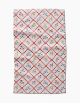 The Fall Flowers tea towel by Claire Iglesias is white with a red diamond lattice and vibrant yellow, red, and blue floral designs inside each diamond. This ultra-absorbent kitchen towel features some visible folds and wrinkles.