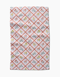 The Fall Flowers tea towel by Claire Iglesias is white with a red diamond lattice and vibrant yellow, red, and blue floral designs inside each diamond. This ultra-absorbent kitchen towel features some visible folds and wrinkles.