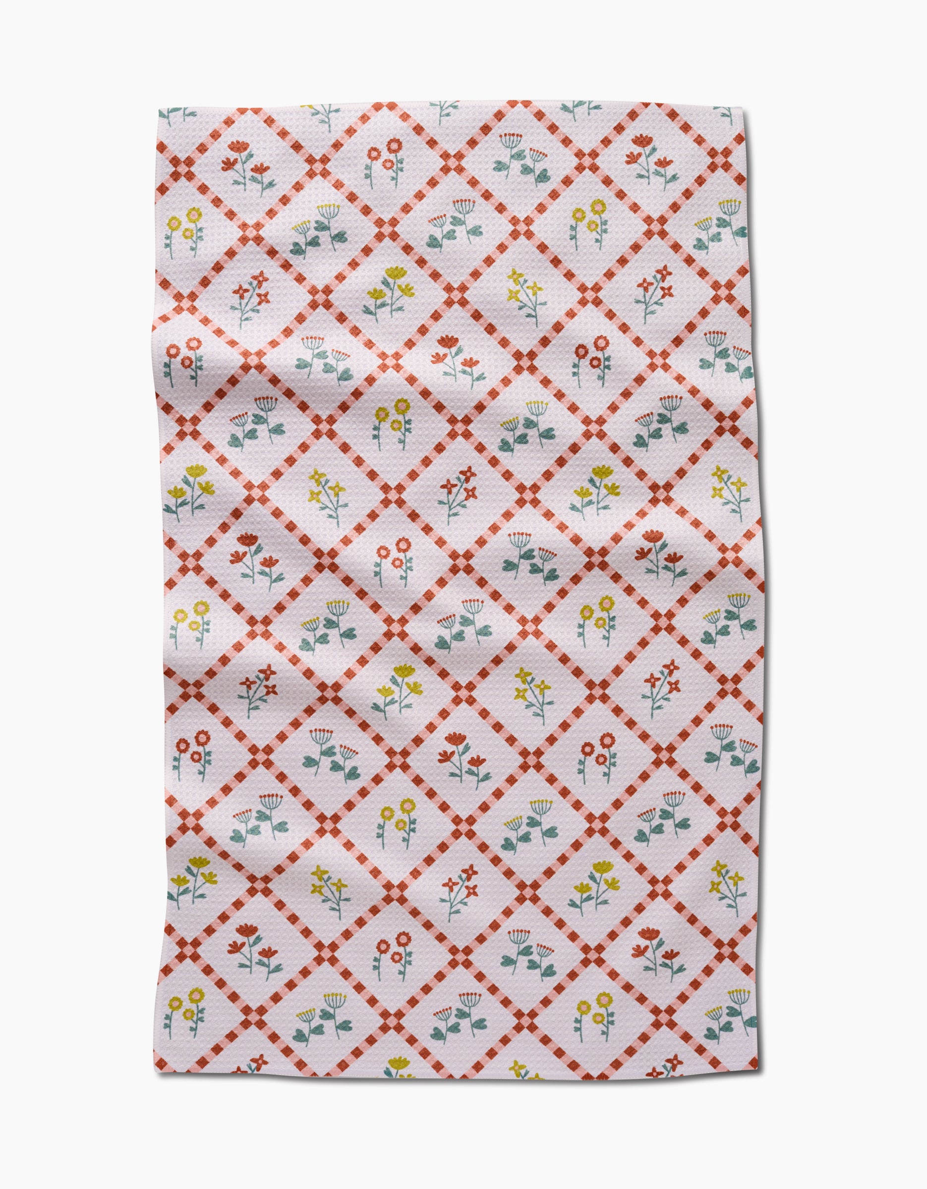 The Fall Flowers tea towel by Claire Iglesias is white with a red diamond lattice and vibrant yellow, red, and blue floral designs inside each diamond. This ultra-absorbent kitchen towel features some visible folds and wrinkles.