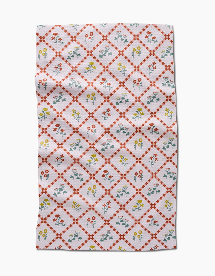 The Fall Flowers tea towel by Claire Iglesias is white with a red diamond lattice and vibrant yellow, red, and blue floral designs inside each diamond. This ultra-absorbent kitchen towel features some visible folds and wrinkles.
